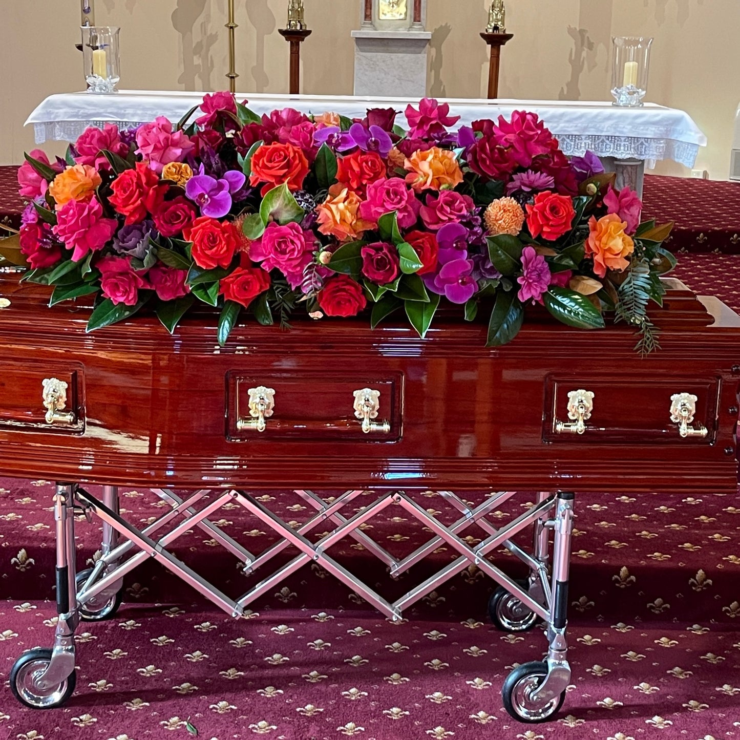 A polished wooden casket with gold handles is elegantly decorated with a Premium Floral Casket Cover from Flowers on Norton St, featuring vibrant red, pink, orange, and purple blooms. It is positioned on a stand within the church, embraced by the tranquil glow of candles and the altar in the background.