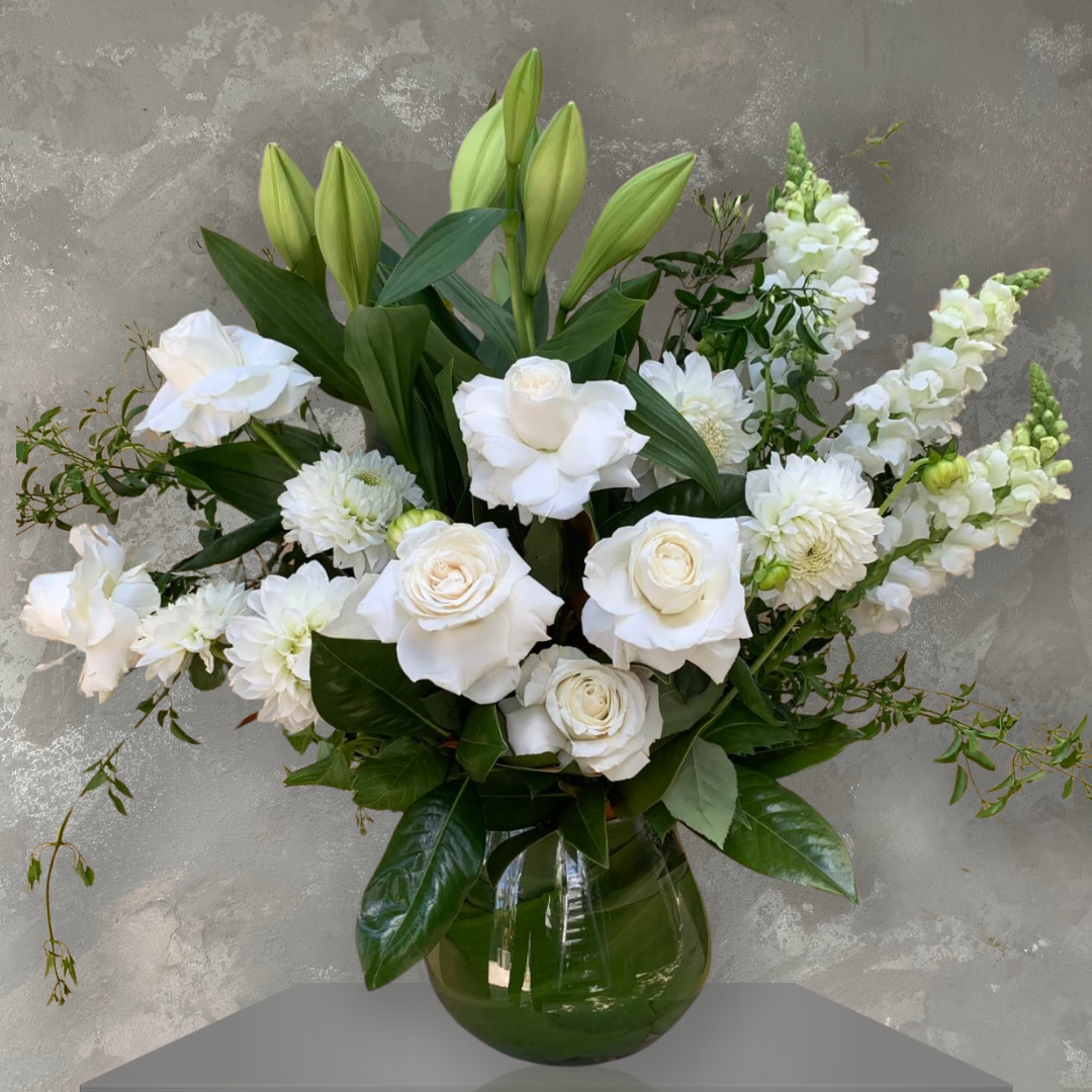 The Celestial Fleurs Sympathy Vase by FlowersonNortonSt features a fishbowl vase filled with a large arrangement of white roses, lilies, and snapdragons, complemented by lush green leaves against a textured gray background—ideal for expressing sympathy.