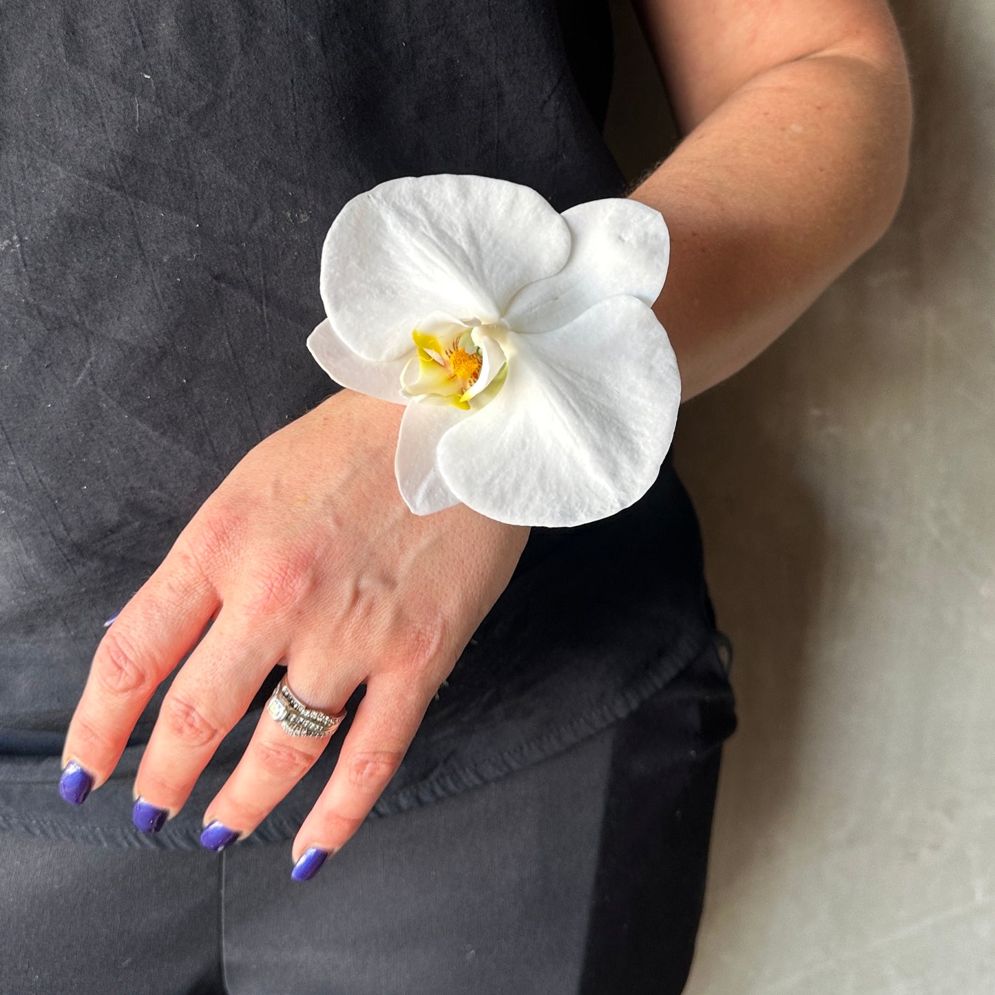 A person dressed in a sleek black ensemble displays their left hand, enhanced with a silver ring and purple nail polish. A stylish Modern Wrist Corsage from Flowers on Norton St elegantly decorates the wrist, featuring a large white orchid that adds an element of sophistication ideal for formal events or school formals.