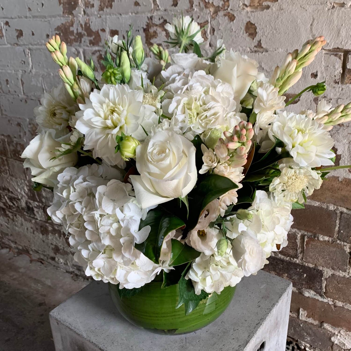 The Divine Fleurs White Flower Vase by Flowers on Norton St showcases a lush bouquet of white roses and hydrangeas in a green vase, displayed on a concrete block against a rustic brick wall.