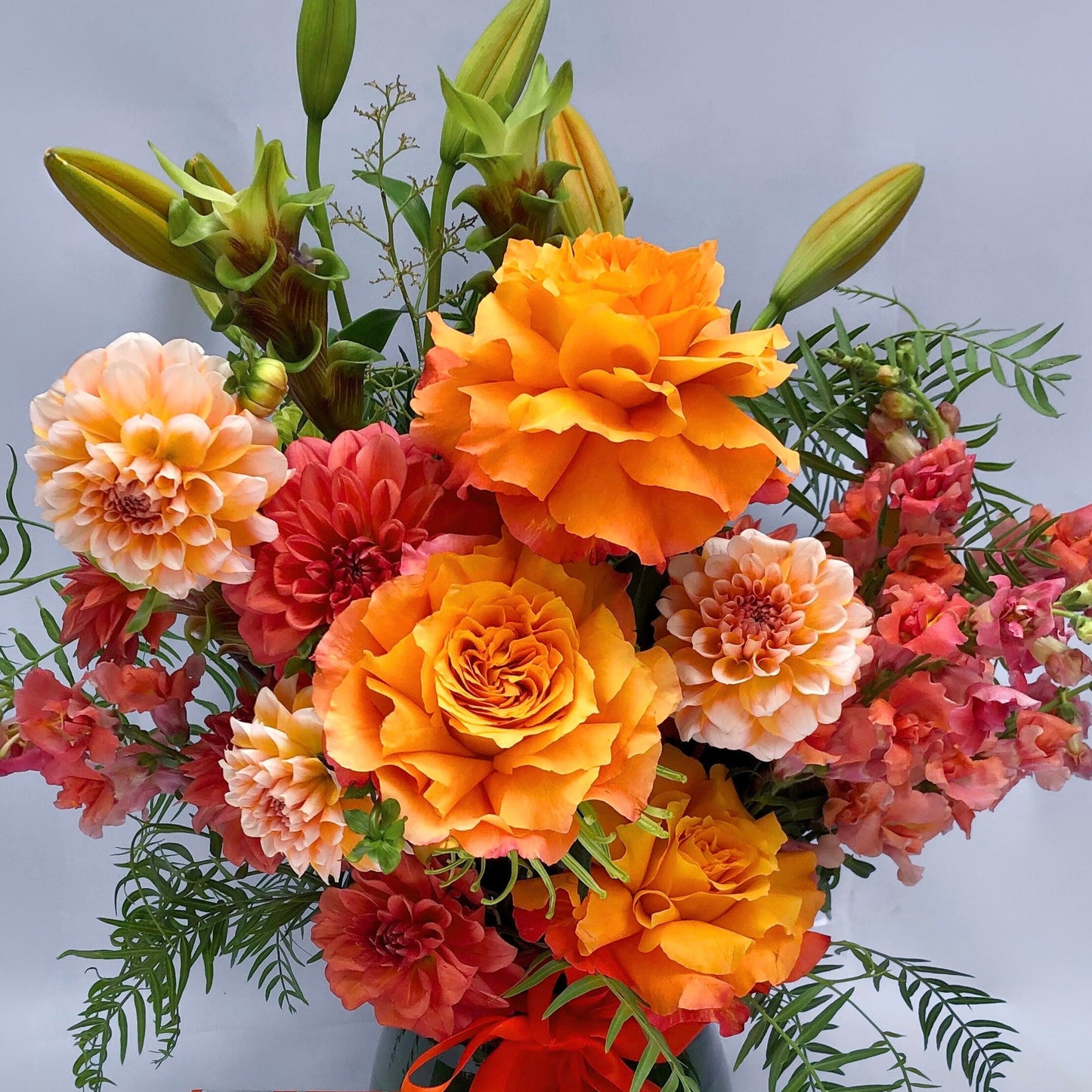 The Tangerine Tango Gift Set by Flowers on Norton St highlights a lively orange floral arrangement featuring peach roses, dahlias, and lilies displayed in a vase with an orange ribbon. The bouquet is complemented by greenery, and it's paired with an orange and white patterned gift box.