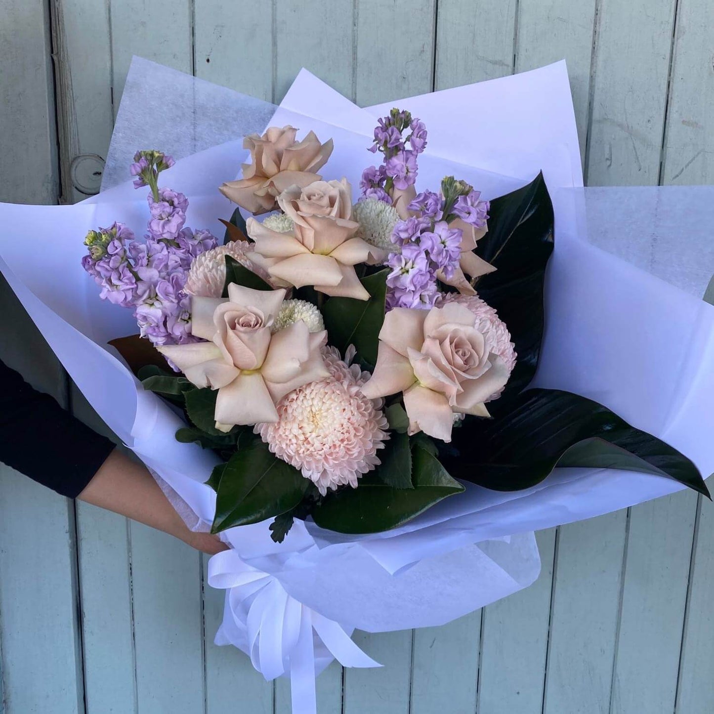 Presenting the "Mauve + White Delight" by Flowers on Norton St: A charming bouquet gift showcasing pale pink roses, light purple stock flowers, and pink chrysanthemums. This delightful arrangement is beautifully enhanced with dark green leaves and skillfully handcrafted by our talented florists. It's elegantly wrapped in white paper and tied with a classic white ribbon.