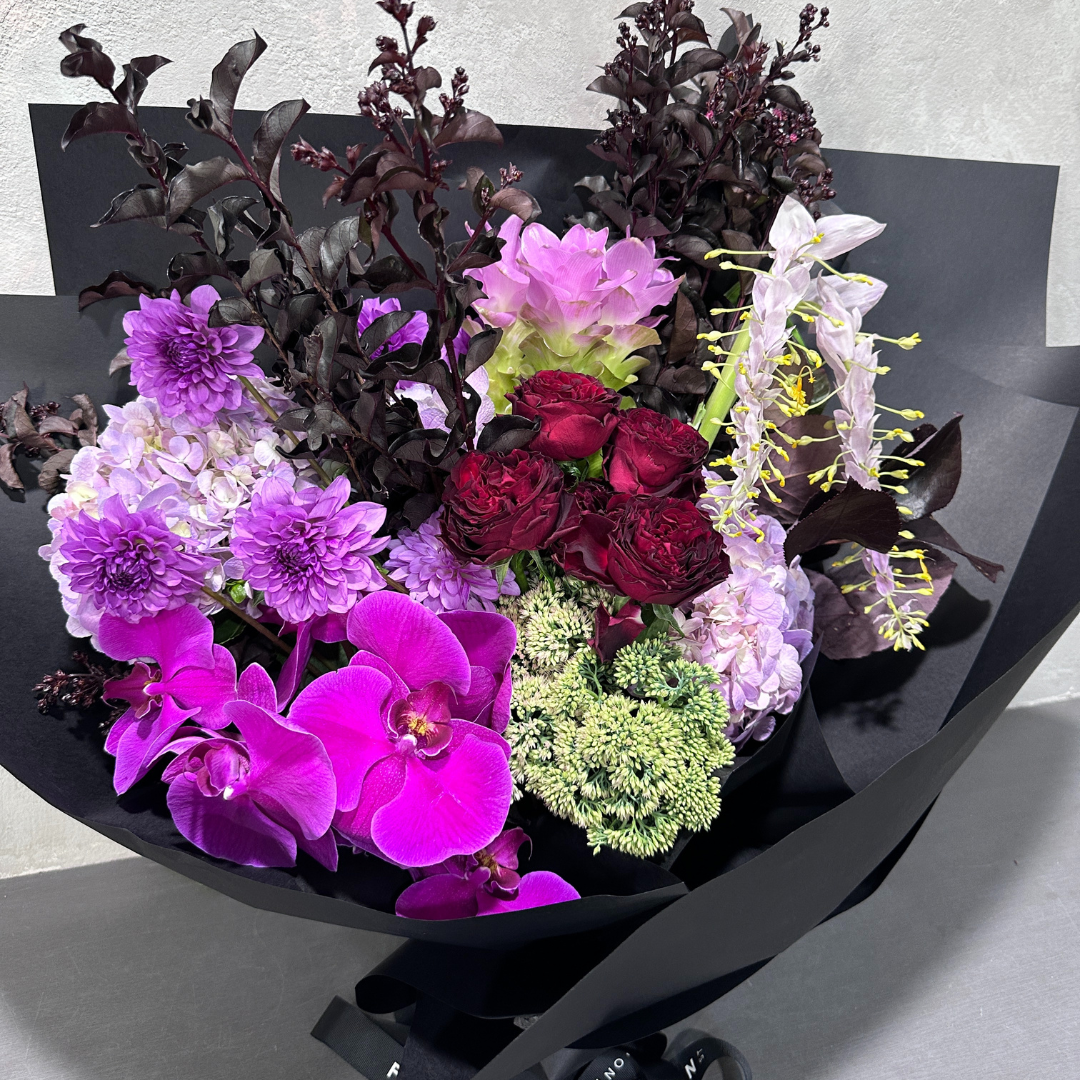 The Purple Delight Bouquet by Flowers on Norton St is a premium-sized, eye-catching display wrapped in black paper. It features vibrant purple dahlias, pink orchids, deep red roses, and assorted greenery set against a neutral background.