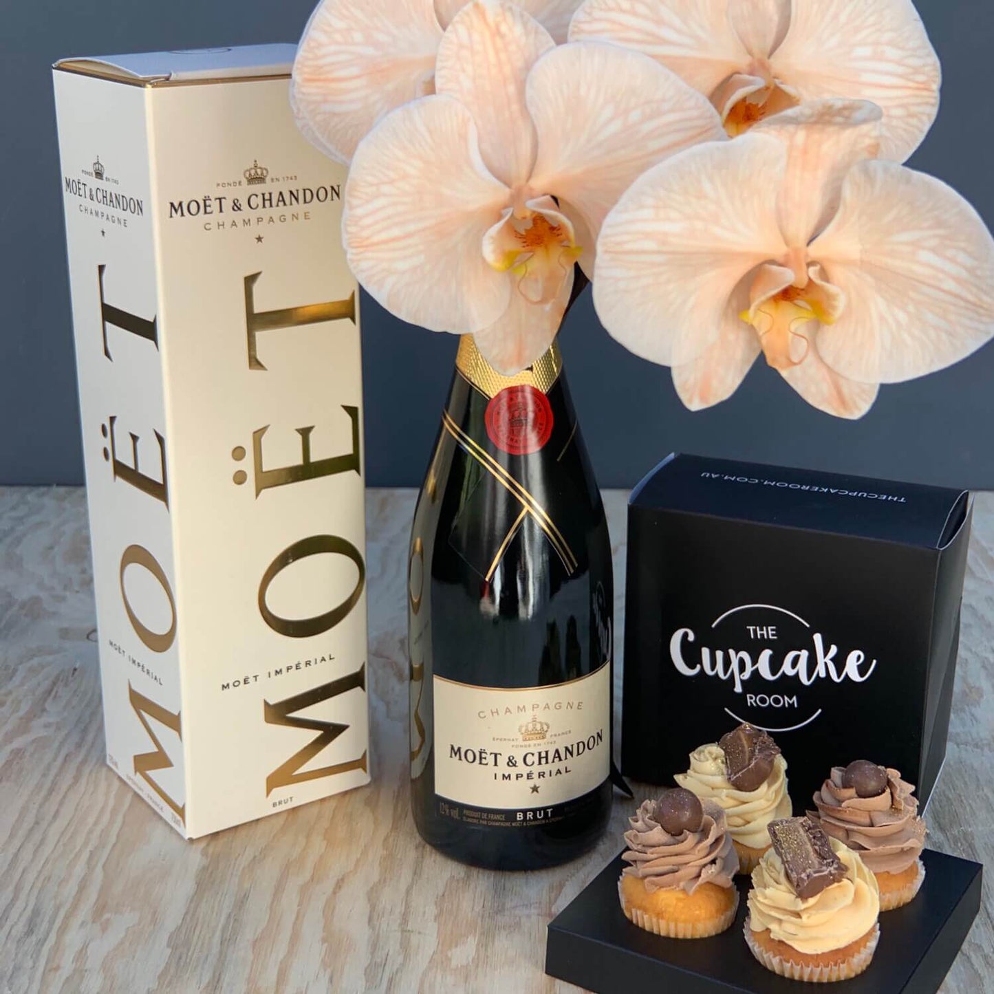 A beautifully styled arrangement titled "Love This Styling" by Flowers on Norton St features a bottle of Moët & Chandon champagne with its packaging, paired with a bouquet of light pink phalaenopsis orchids. Accompanying this elegant display is a box from "The Cupcake Room" containing four assorted cupcakes adorned with unique frosting designs, all elegantly presented on a wooden surface.