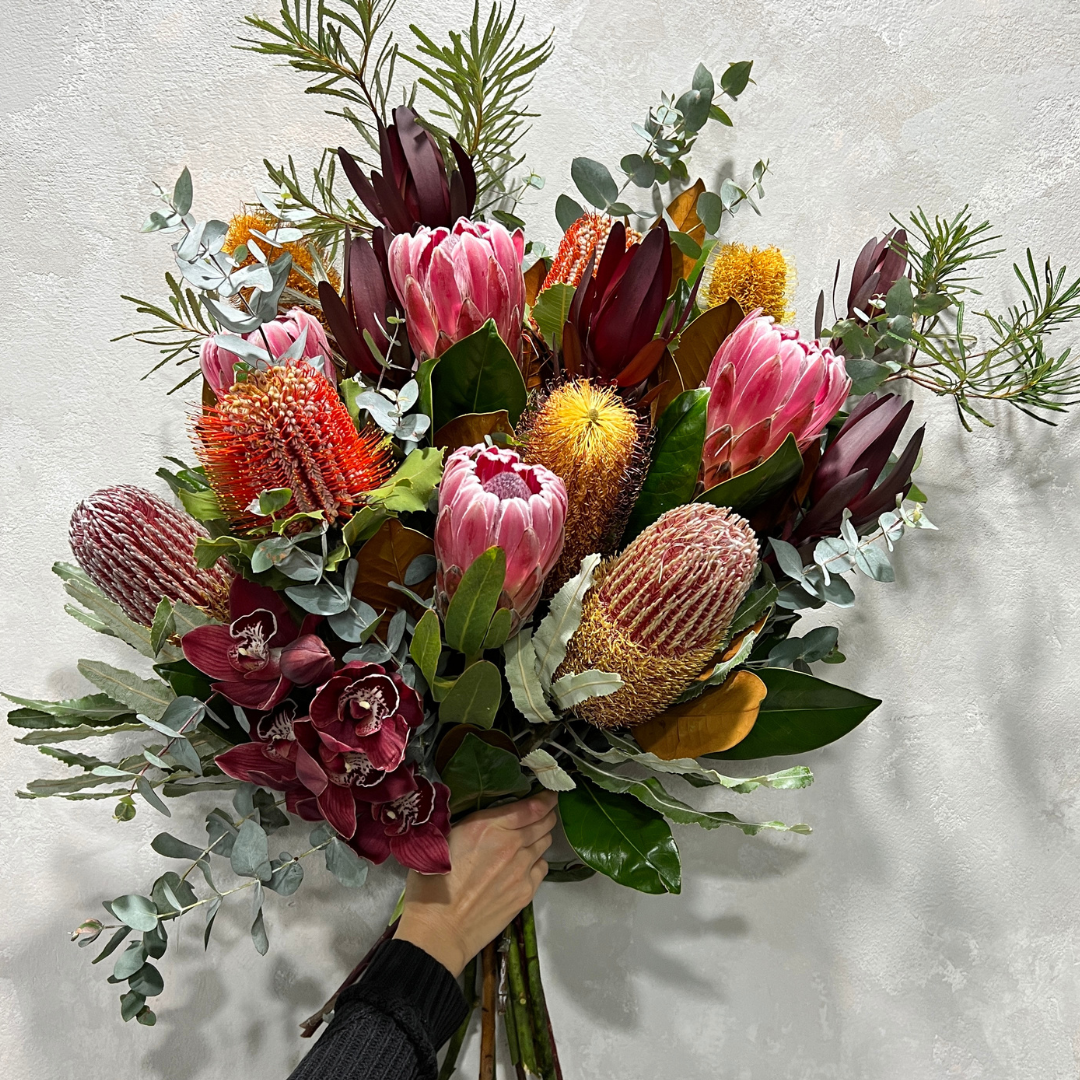 Introducing the "Australiana" by FlowersonNortonSt—a stunning bouquet showcasing an array of Australian wildflowers. This vibrant arrangement features pink proteas, orange and yellow banksias, and red leucadendrons, beautifully intertwined with orchids and eucalyptus, all complemented by lush greenery set against a light background.