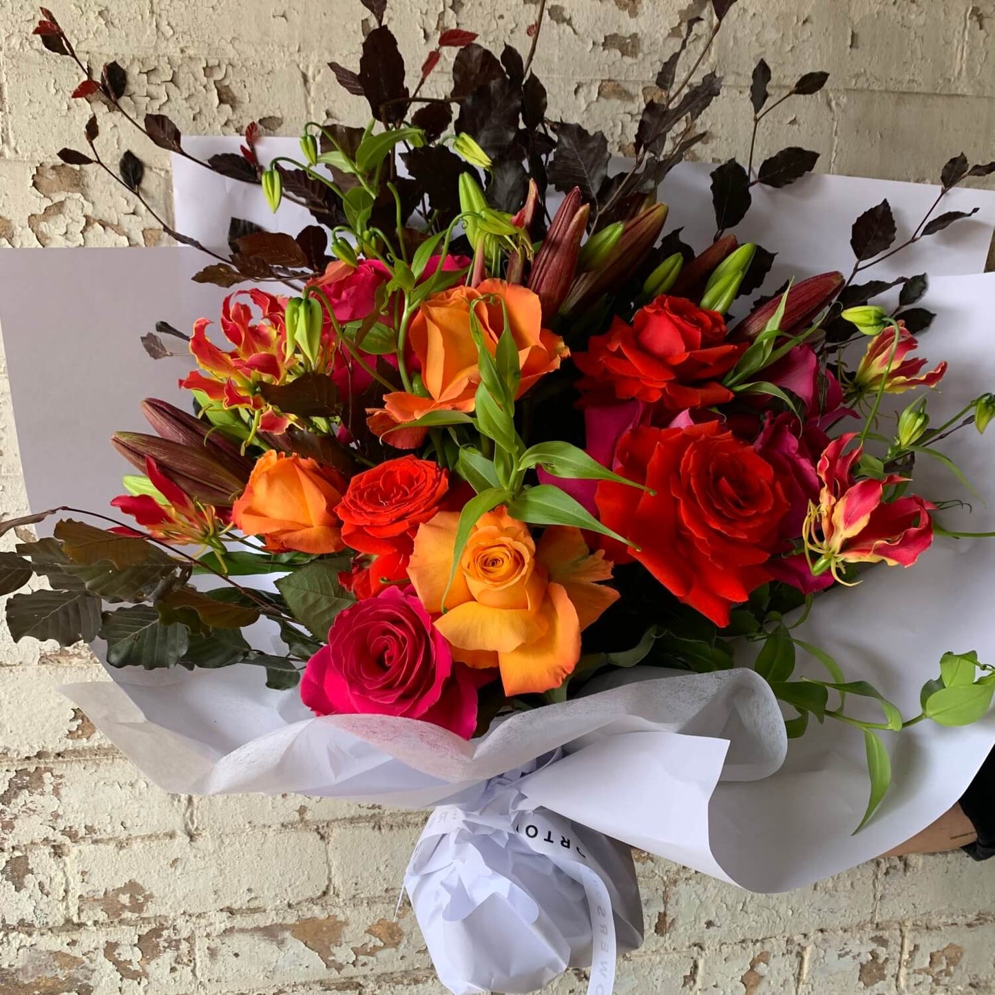 The Floral Decadence Mixed Flower Bouquet by Flowers on Norton St offers a luxurious assortment of red, orange roses, pink blooms, and dark foliage in white wrapping against a rustic brick wall. Ideal for flower delivery in Sydney.