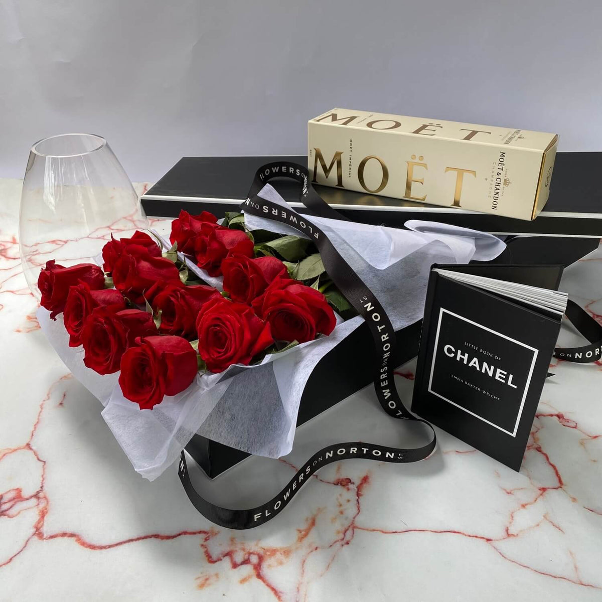 The French Luxury Roses Gift Set by FlowersonNortonSt includes red roses in white paper, a Moët champagne box with a glass, and a Chanel book on marble. Black gift boxes with ribbons complete this luxe scene.