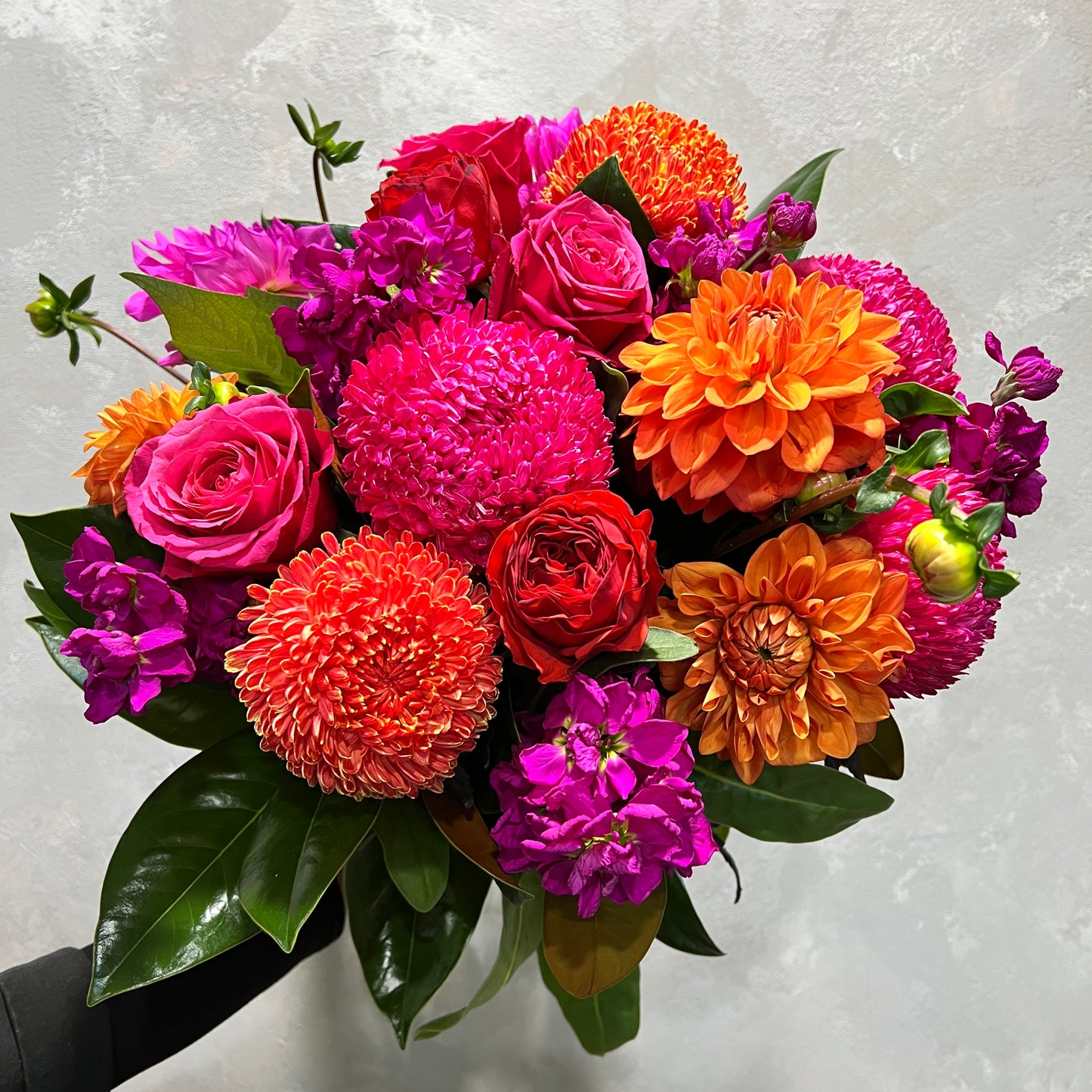 The Summertime Pink + Orange Mixed Flowers from FlowersonNortonSt is a celebratory bouquet that features radiant orange and pink flowers, including roses, chrysanthemums, and dahlias, accented with lush green leaves. This vibrant arrangement evokes the hues of summer sunsets against a light gray background.