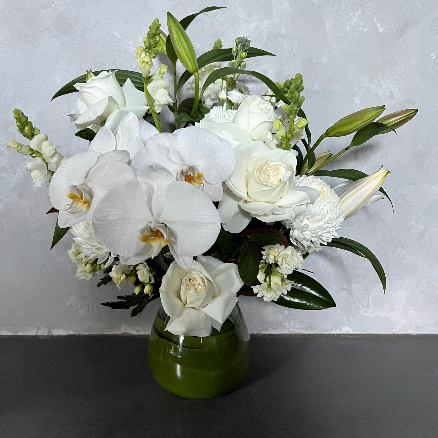 The "Sympathy Mixed Flower Vase White" by FlowersonNortonSt features a stylish bouquet of white blooms, including orchids, roses, lilies, and snapdragons elegantly arranged in a green glass vase set against a light gray textured background. It's perfect for expressing condolences with grace and beauty.