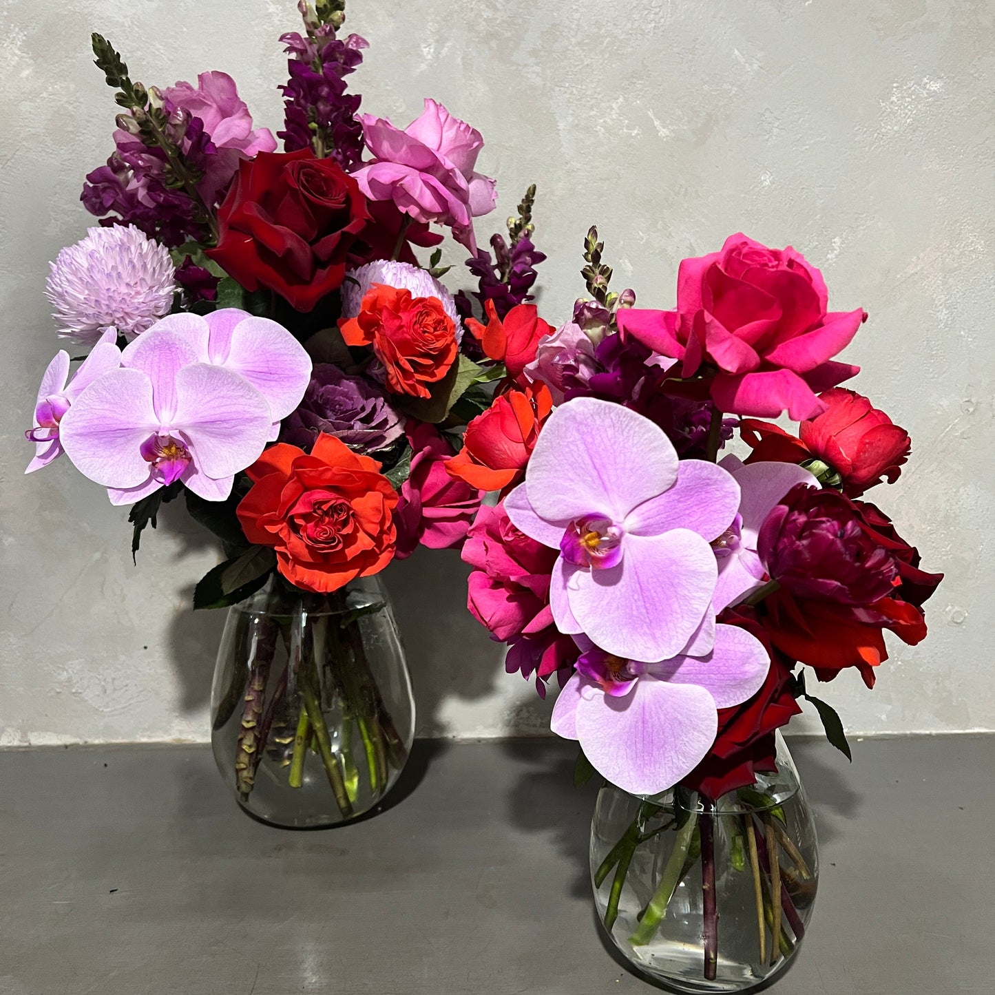 Two Berry Expression Mixed Flower Vases from FlowersonNortonSt display vibrant flower arrangements against a neutral backdrop. Each teardrop vase showcases a colorful and lush mix of pink orchids, red roses, and seasonal purple blooms.