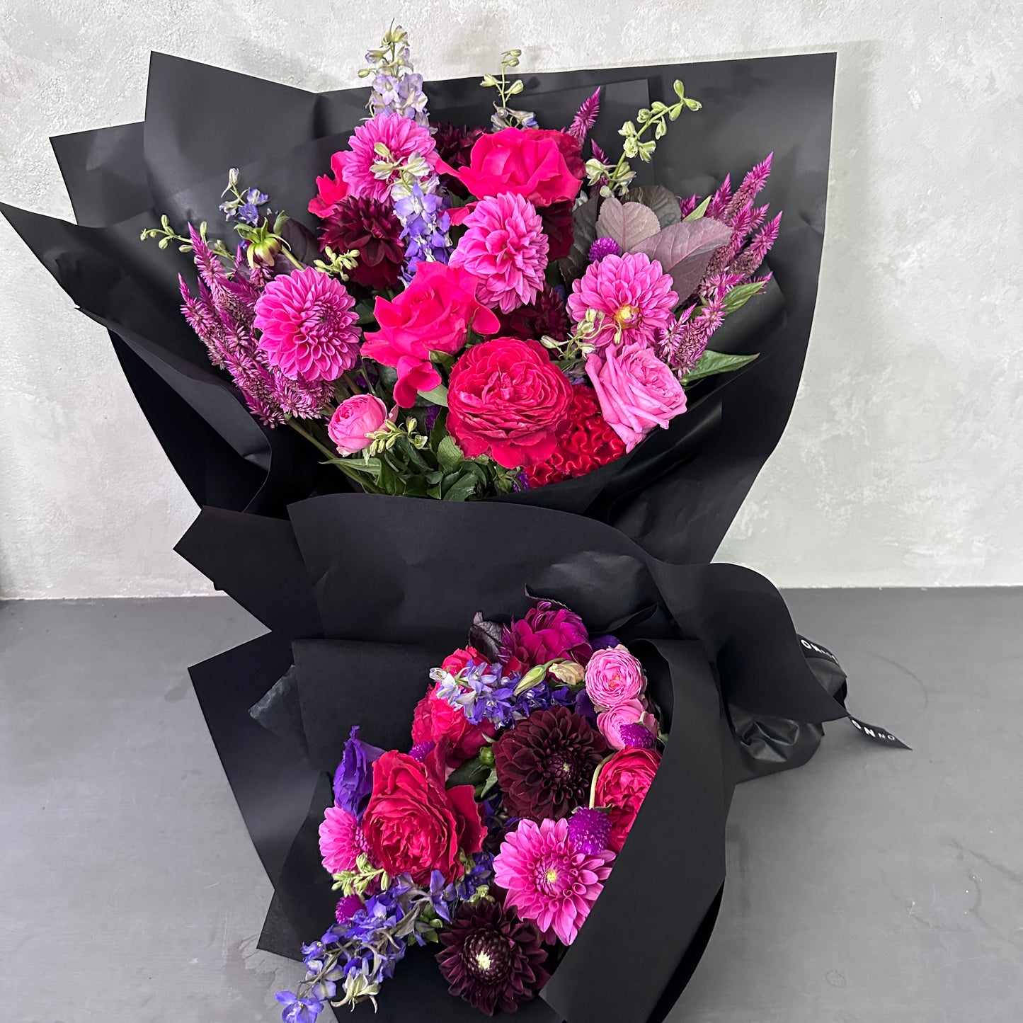 Product from Flowers on Norton St: The Hot Pink Pop Mixed Flower Bouquet showcases two elegant arrangements wrapped in black paper, featuring vibrant seasonal blooms in shades of pink, purple, and red. These bouquets include dahlias, roses, and assorted greenery for a modern and sophisticated floral display against a subtle gray background.
