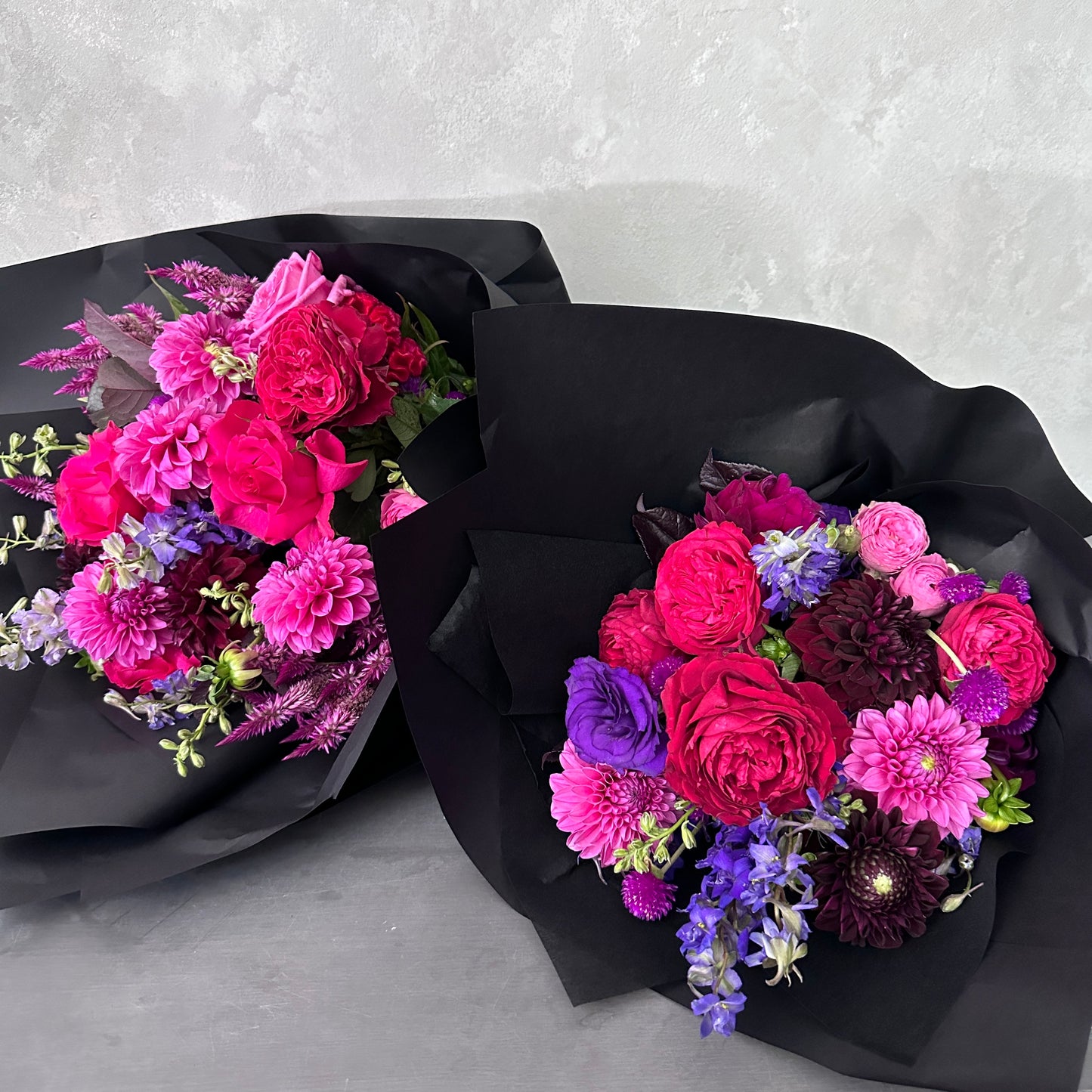 The Hot Pink Pop Mixed Flower Bouquet by Flowers on Norton St features two elegant floral arrangements wrapped in black paper. Each bouquet showcases a vibrant mix of fuchsia, pink, and purple flowers, including roses and dahlias, artfully crafted from seasonal blooms against a light gray background.