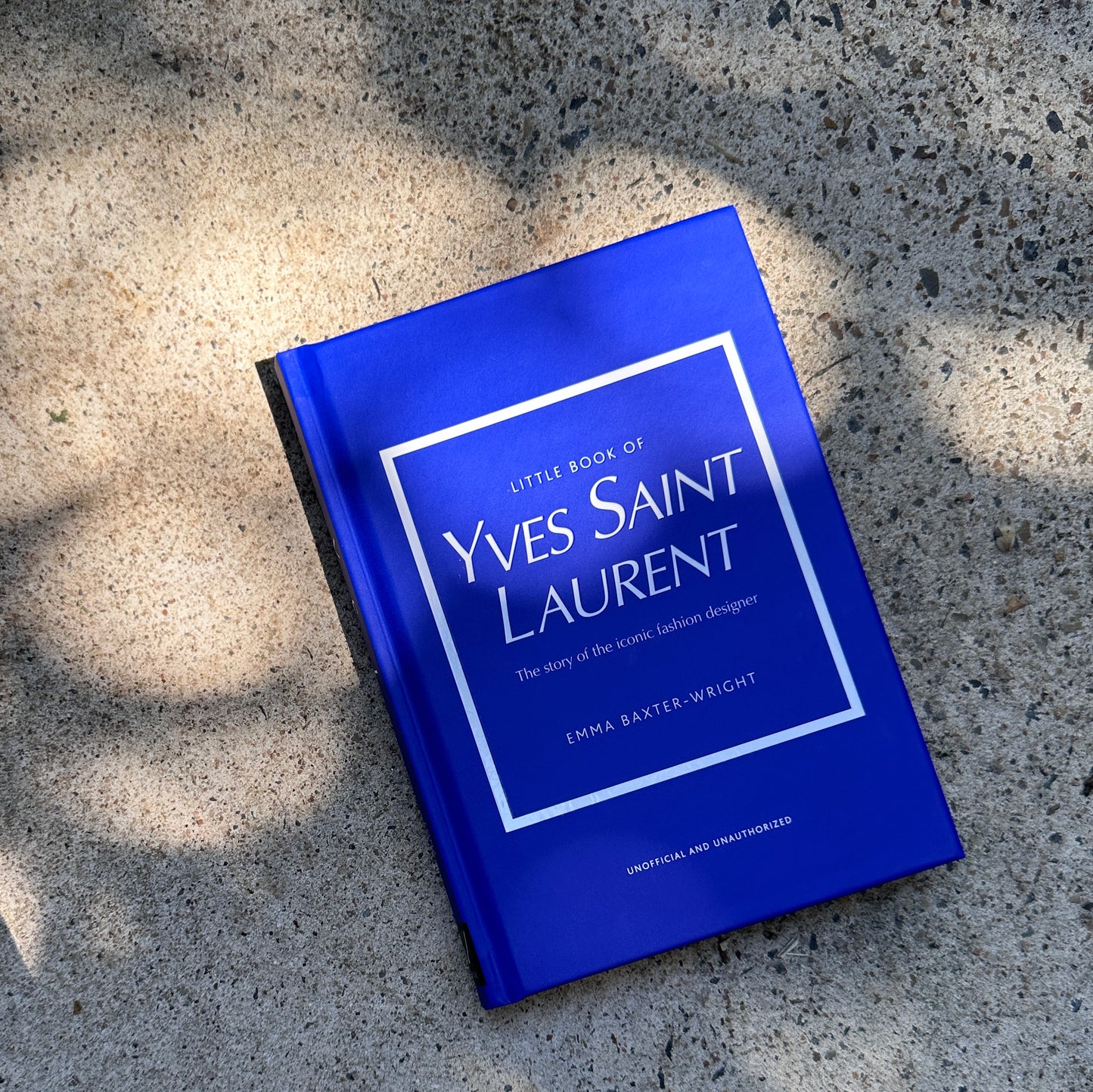 A book with a blue cover titled "Little Book of Style" by Flowers on Norton St, ideal for anyone passionate about fashion, is placed on a textured stone surface where gentle shadows gracefully drape over it. As part of the "Little Guides to Style," it honors legendary fashion houses.