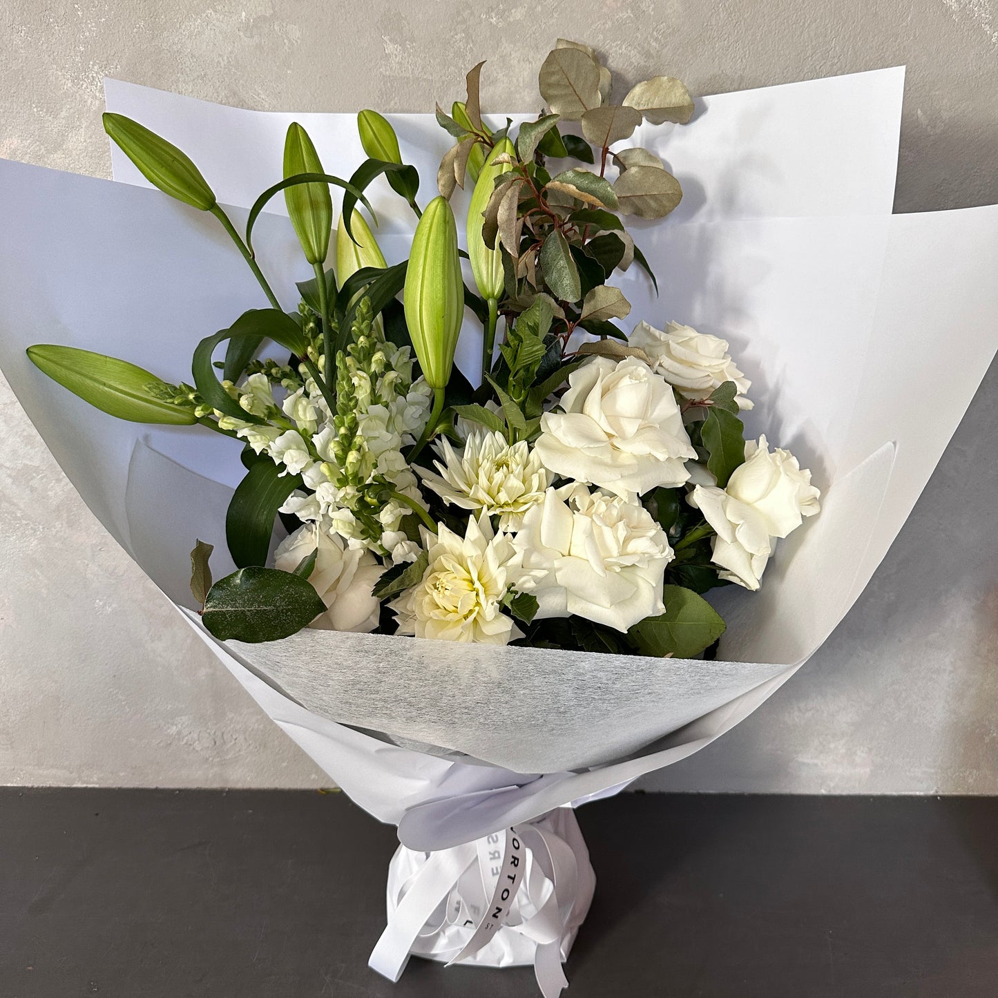 Introducing the Cream Classic Mixed Flowers by FlowersonNortonSt: a stunning arrangement of white and cream blooms, featuring roses and lilies, surrounded by statement greenery and wrapped in elegant white paper with a delicate white ribbon. This sophisticated selection is beautifully presented against a light gray background.