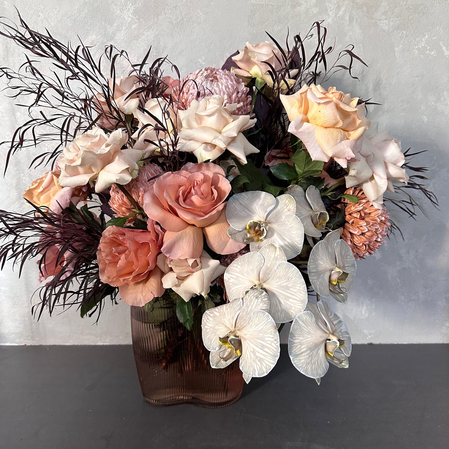 The Peaches + Cream Mixed Flower Vase by Flowers on Norton St features Colombian roses, Cloud Moth orchids, peach and cream roses, pink dahlias, white orchids, and dark wispy foliage in a textured brown vase. It beautifully contrasts a light gray background.