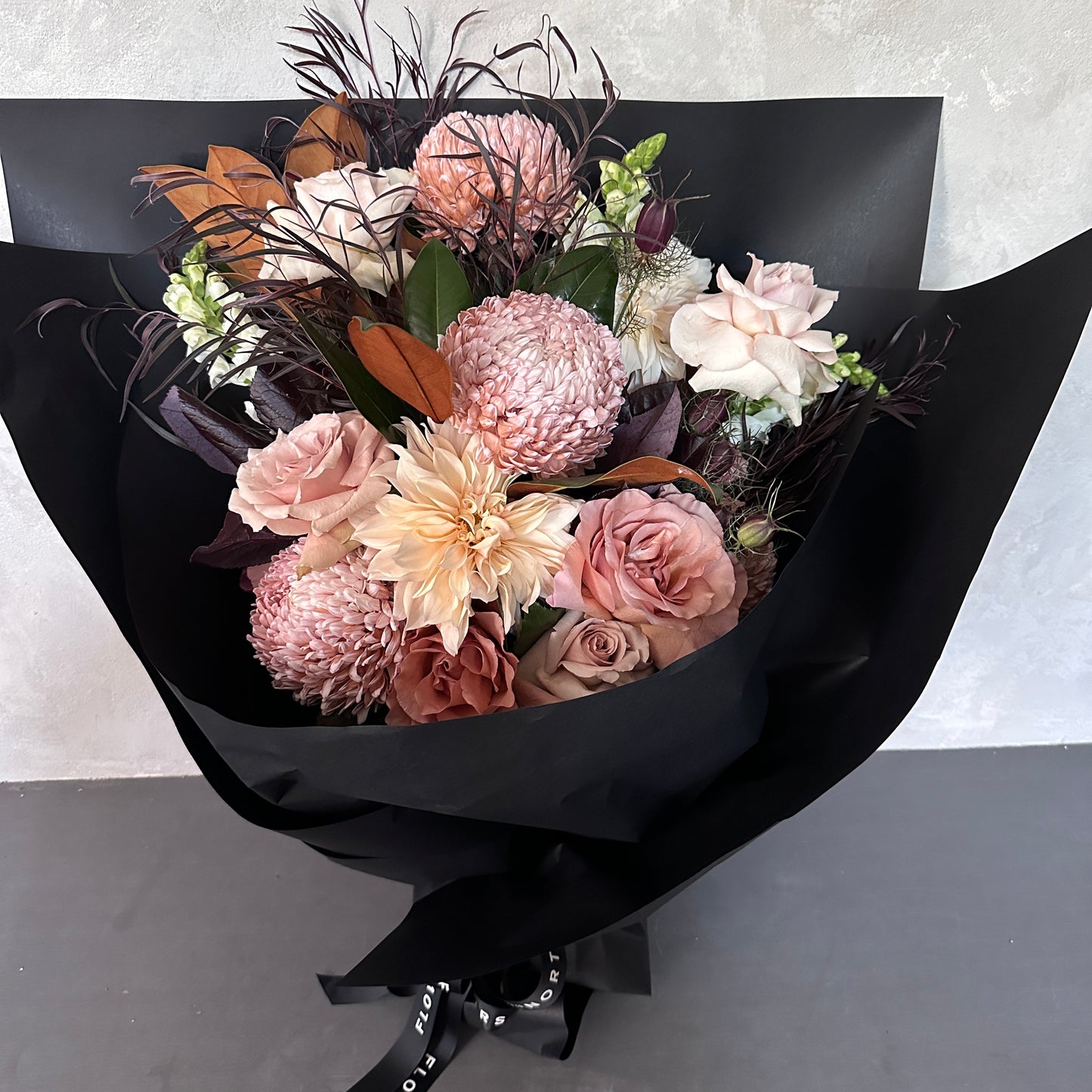 The "Peaches & Cream" bouquet from Flowers on Norton St features luxury flowers in pink and peach hues, elegantly wrapped in black paper. This arrangement includes Colombian roses and chrysanthemums, accented with leaves, beautifully presented against a light gray background.