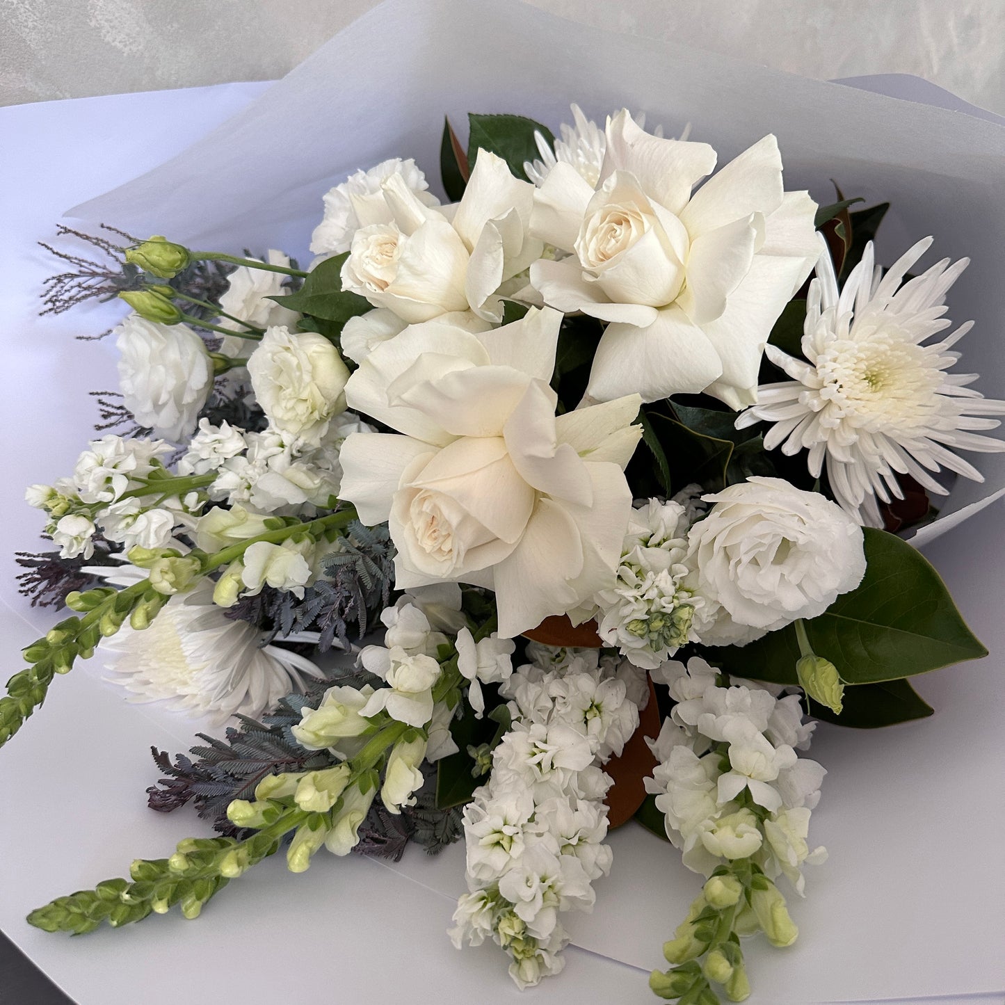 The Serenity Sheath by FlowersonNortonSt is a carefully arranged floral sheaf featuring white roses, mums, and snapdragons complemented by green leaves, all elegantly wrapped in white paper—an ideal tribute for offering sympathy.