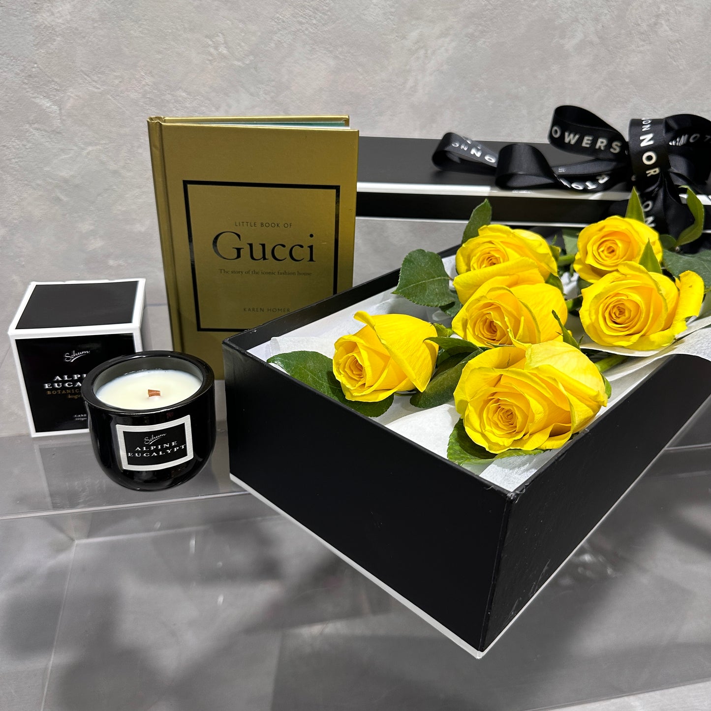 A chic arrangement features FlowersonNortonSt's The Rose Gift Box filled with yellow roses, an open book titled "Little Book of Gucci," a black candle named "Alpha Escape," and a black-and-white ribbon, all elegantly displayed on a clear surface against a gray backdrop. This sophisticated setup creates an unforgettable gift.