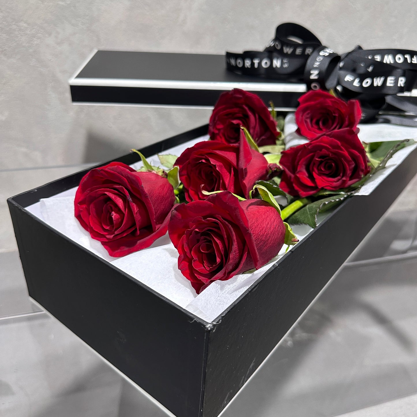 The Classic Rose Box by FlowersonNortonSt offers a memorable gift, featuring six partially open red roses arranged on white paper to enhance their vibrant beauty. The sleek design of the box is elegantly wrapped with a black ribbon featuring white text.