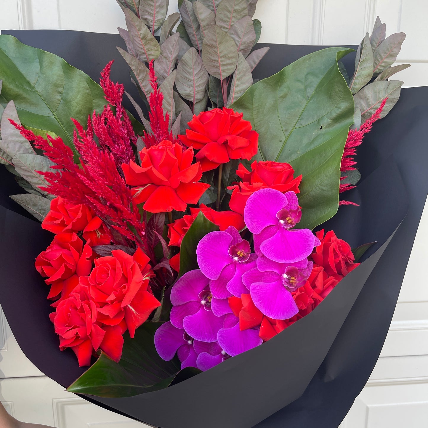 The Strawberry Spritz Red + Magenta Bouquet by FlowersonNortonSt showcases Nina roses and purple Phalaenopsis orchids nestled in verdant foliage, all beautifully wrapped in black paper against a neutral background.