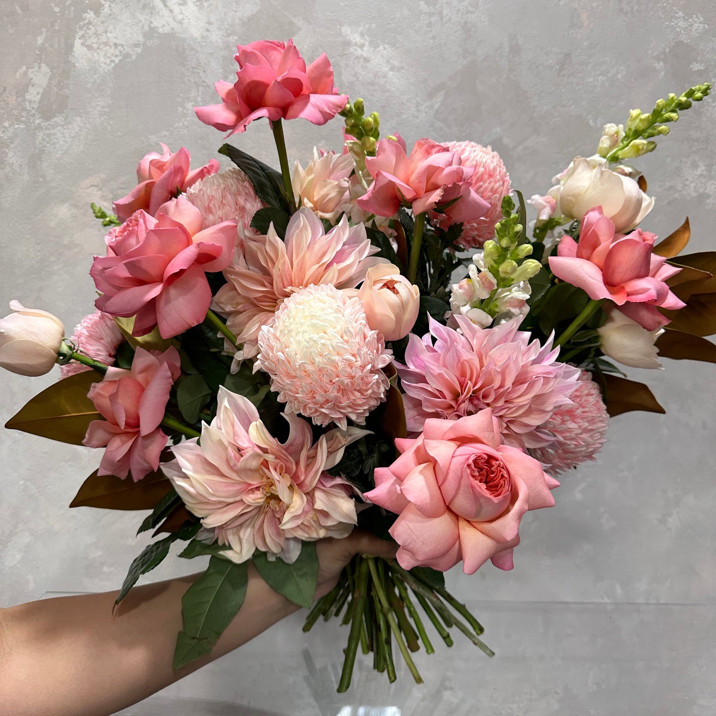 The Pink Expression Seasonal Bouquet 2024 by Flowers on Norton St is a vibrant mix of dahlias, roses, and snapdragons with green leafy accents and seasonal blooms set against a textured gray backdrop.
