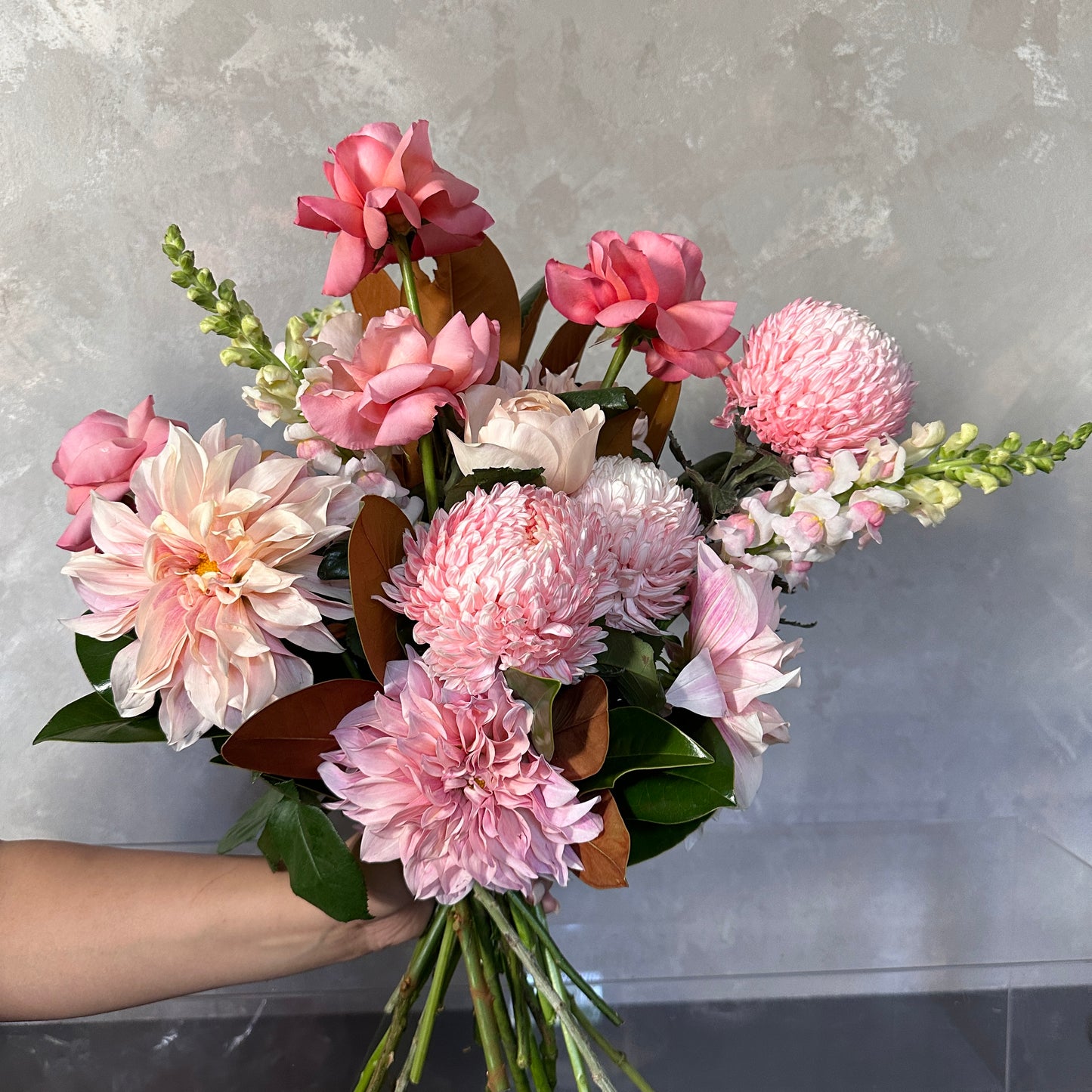 A hand holds the Pink Expression Seasonal Bouquet 2024 by Flowers on Norton St, featuring pink roses, dahlias, and seasonal blooms with lush green leaves, set against a textured gray background.