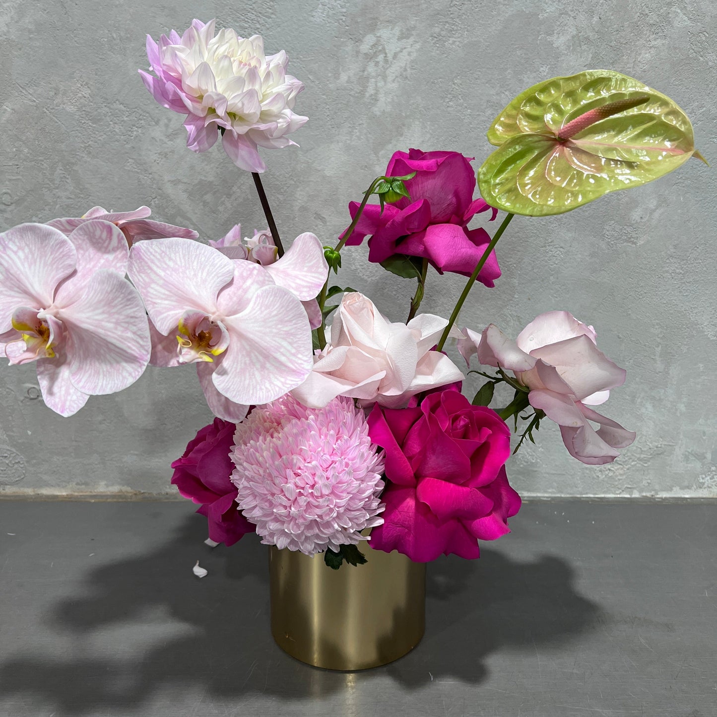 The "Pink Pop" by Flowers on Norton St presents a modern composition in a gold vase, highlighting white and pink orchids, pink roses, a chrysanthemum, and green anthurium against a light gray textured background.