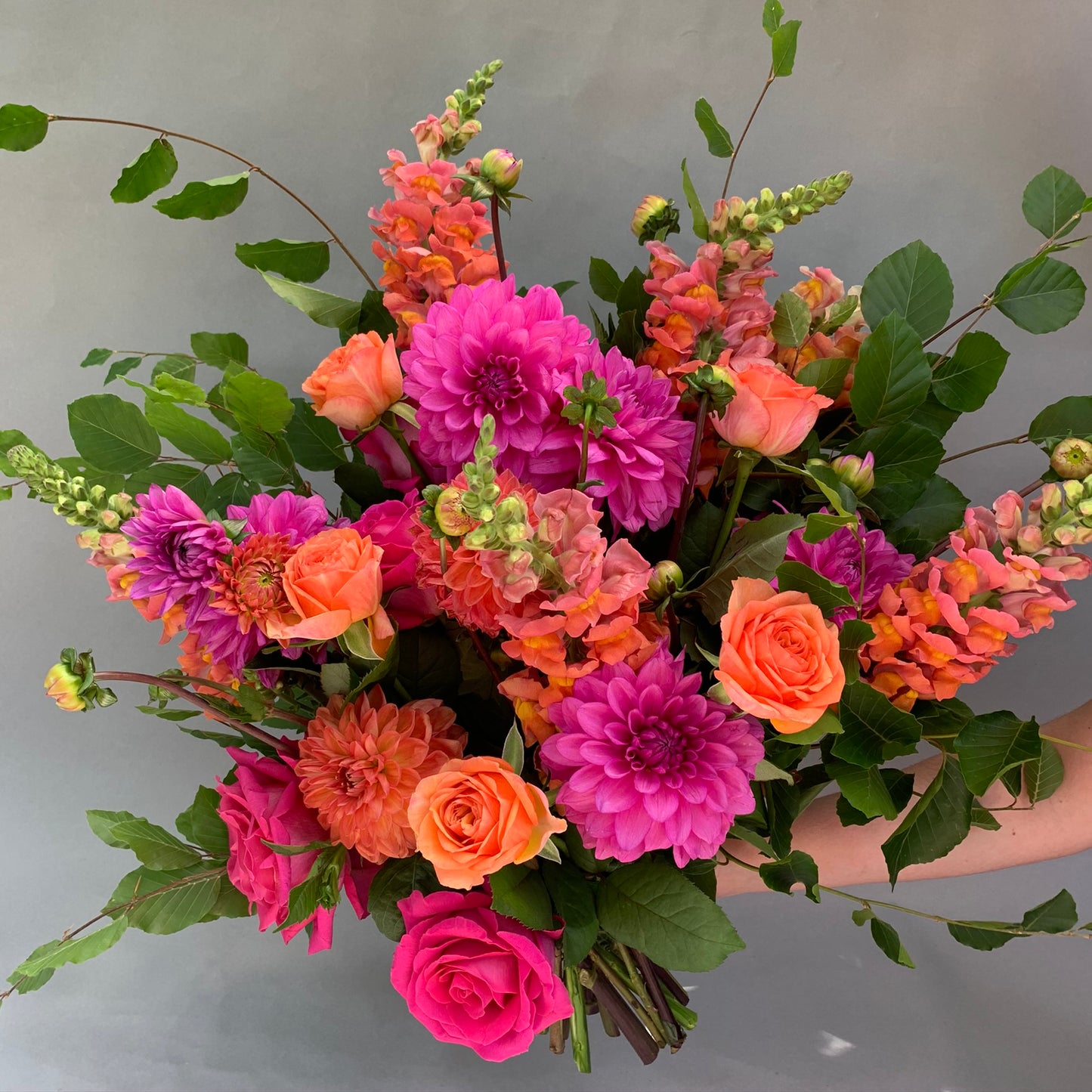 Introducing the Summertime Pink + Orange Mixed Flowers by FlowersonNortonSt, a delightful bouquet featuring pink dahlias, orange roses, and multicolored snapdragons nestled among lush green leaves, evoking the charm of summer sunsets against a gray backdrop.