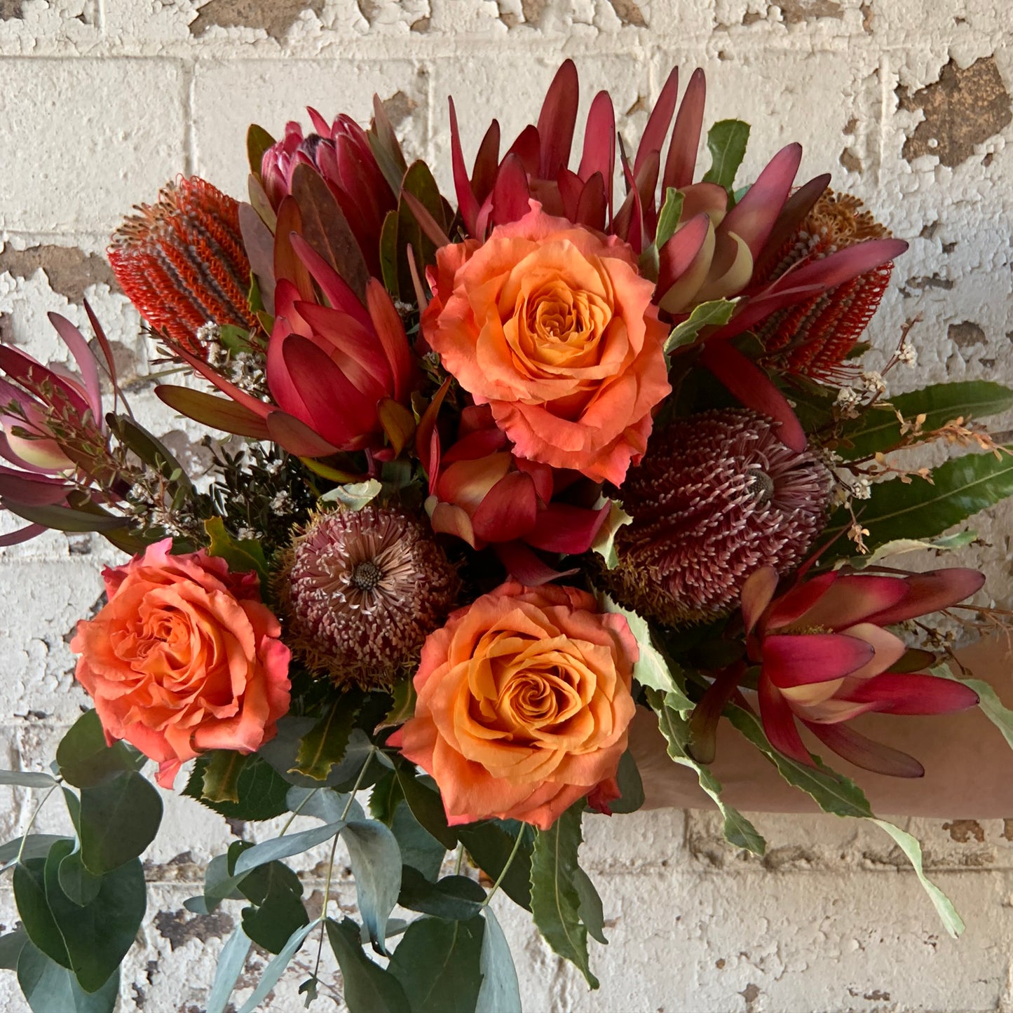 The Eora Sunset Natives + Rose Bouquet by FlowersonNortonSt is a vibrant floral arrangement featuring burnt orange roses and red protea, highlighted with native wildflowers and assorted greenery against a textured white brick wall. This combination of colors and textures creates a striking, warm display that offers a long-lasting flower choice.