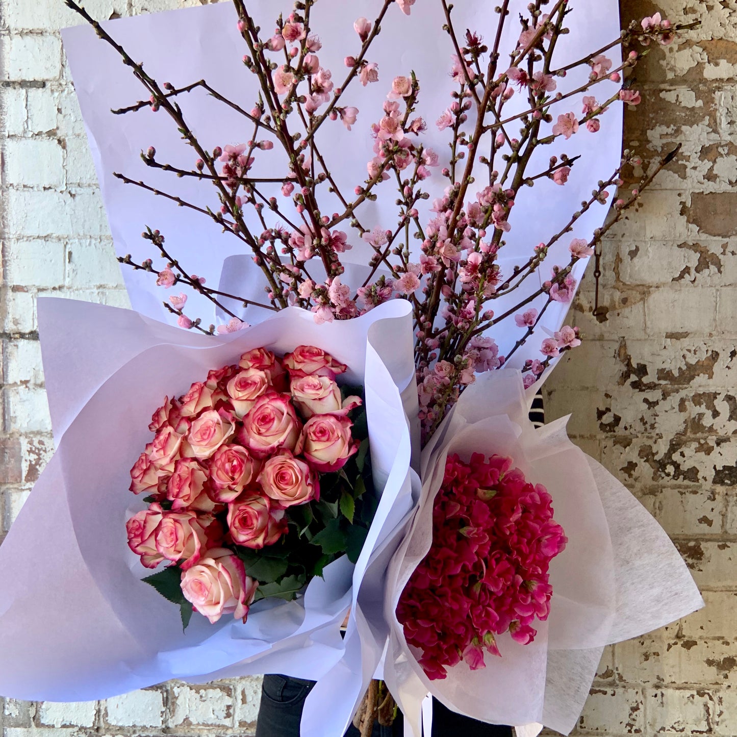 The "Colour Me Pink Seasonal Flowers" arrangement by Flowers on Norton St features peach blossoms at the center, flanked by a bouquet of seasonal pink and white roses on one side and bright pink hydrangeas on the other, all elegantly wrapped in white paper set against a textured brick wall.