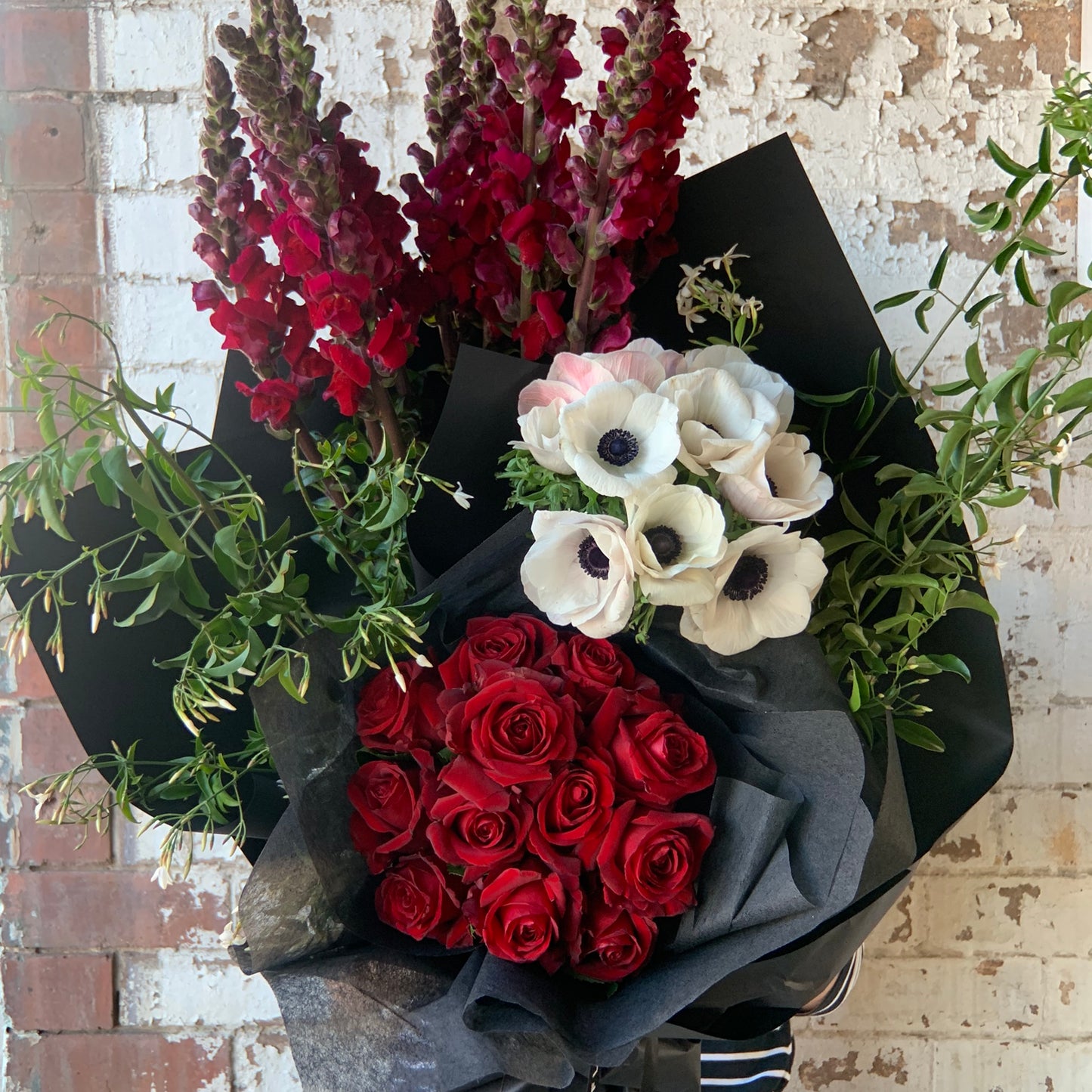 The "Colour Me - Red Seasonal Flowers" bouquet by Flowers on Norton St makes a romantic gift with its deep red roses, white anemones featuring dark centers, and tall dark red snapdragons. These beautiful seasonal flowers are complemented by green foliage and elegantly wrapped in black paper, set against a brick wall backdrop.