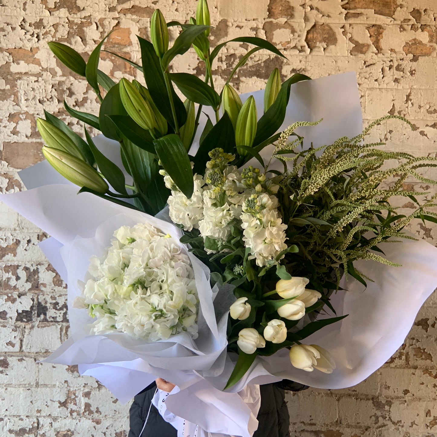 The "White Delight Seasonal Flowers" bouquet by FlowersonNortonSt, which includes white lilies, hydrangeas, and tulips elegantly wrapped in white paper, is perfectly complemented by the rustic white brick wall backdrop. This setting highlights the elegant contrast of lush green leaves with delicate seasonal blooms.