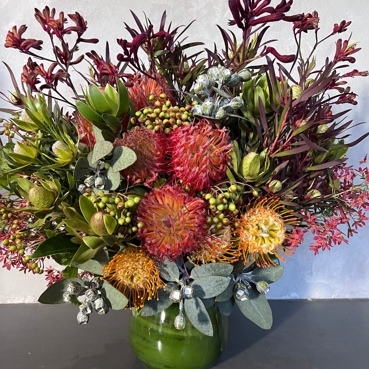 Presenting the Wangal Australian Natives Vase Bouquet by FlowersonNortonSt: a lively arrangement showcasing red and orange pincushion proteas paired with lush tropical foliage, complemented by small clusters of berries in a glossy green vase. This sculptural vase bouquet is beautifully set against a light, textured background, adding an artistic flair.