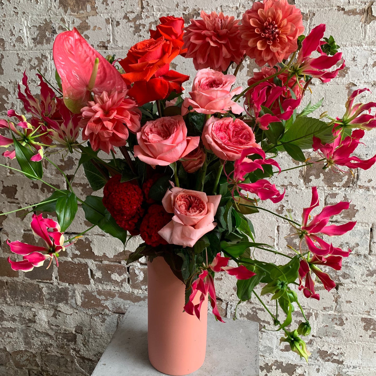 The Summer Splice arrangement by FlowersonNortonSt beautifully displays flame lilies, red and pink roses, and seasonal dahlias in a pink vase against a textured brick wall—ideal for housewarming occasions.