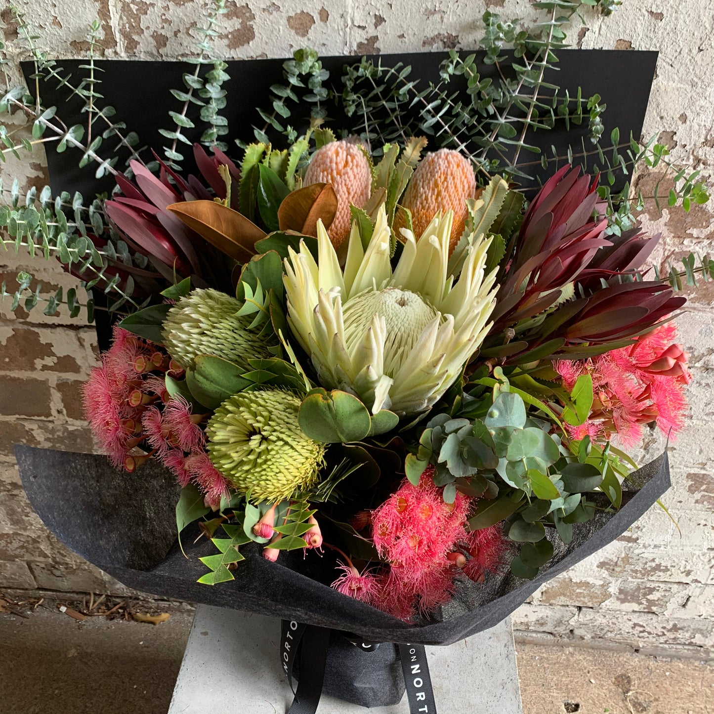 The Native Flare Bouquet by FlowersonNortonSt features a stunning large white protea at its center, surrounded by colorful pink, orange, and green Australian native flowers accompanied by eucalyptus leaves. This vibrant wildflower arrangement is beautifully showcased against a rustic brick wall backdrop.