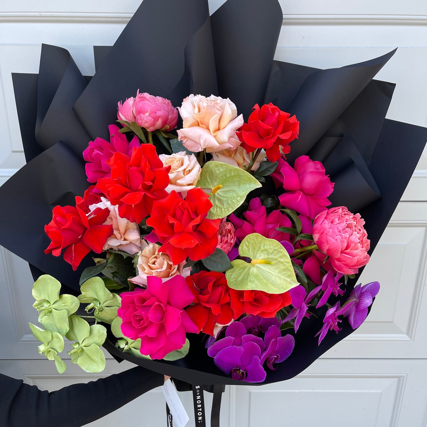 The Hello Lovely Rose + Anthurium Bouquet by FlowersonNortonSt showcases a vibrant assortment of flowers, including flexed roses in red, pink, and peach hues. These are elegantly surrounded by lush green leaves and beautifully complemented by purple orchids, all wrapped in chic black paper for a stunning botanical display.