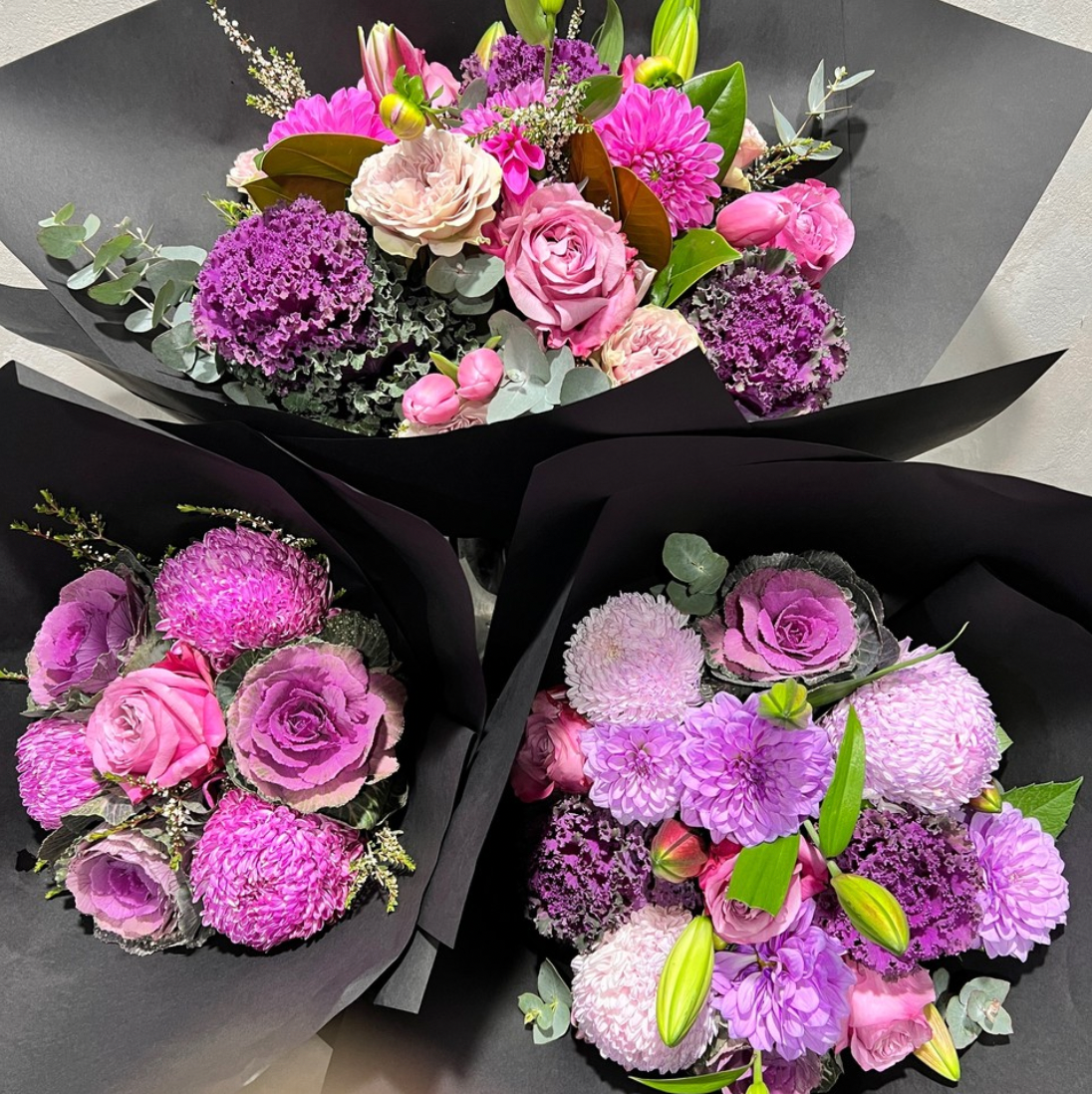 Three "Not Over The Top Seasonal Bouquets" by FlowersonNortonSt feature pink and purple flowers wrapped in black paper. Each arrangement includes seasonal blooms like roses, dahlias, and greenery, creating a vibrant and elegant display.