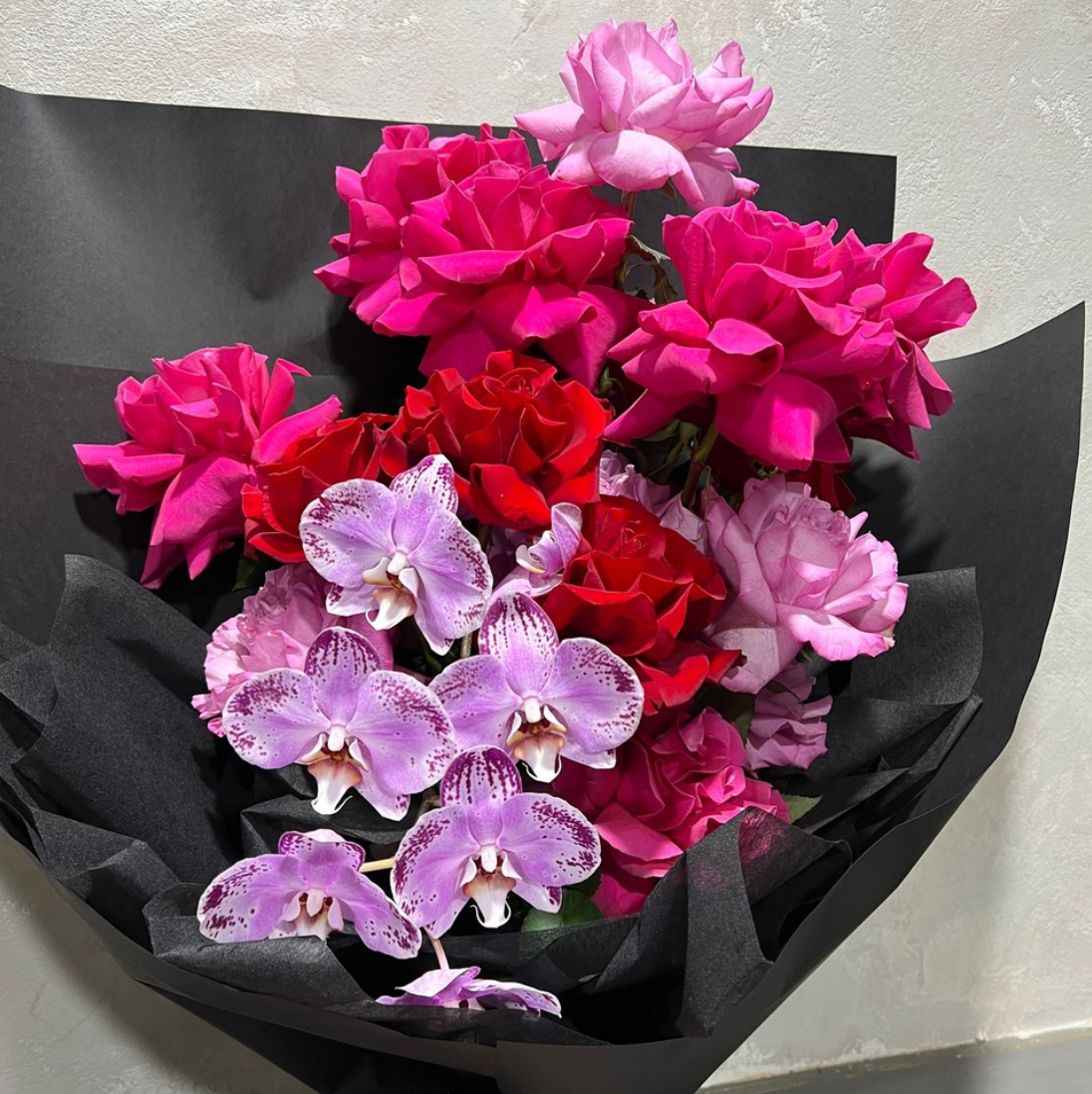 The Flamenco Fusion Bouquet by FlowersonNortonSt showcases vibrant pink and red roses, paired with purple orchids accented by white speckles, all elegantly wrapped in a stylish ruffle wrap against a plain, light-colored background.