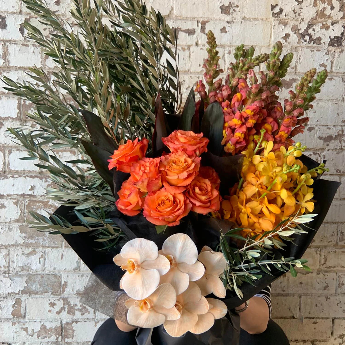The Orange Crush Seasonal Bouquet by Flowers on Norton St showcases a vibrant mix of orange roses, yellow orchids, pink snapdragons, and white anthuriums with lush green foliage against a rustic white brick wall.