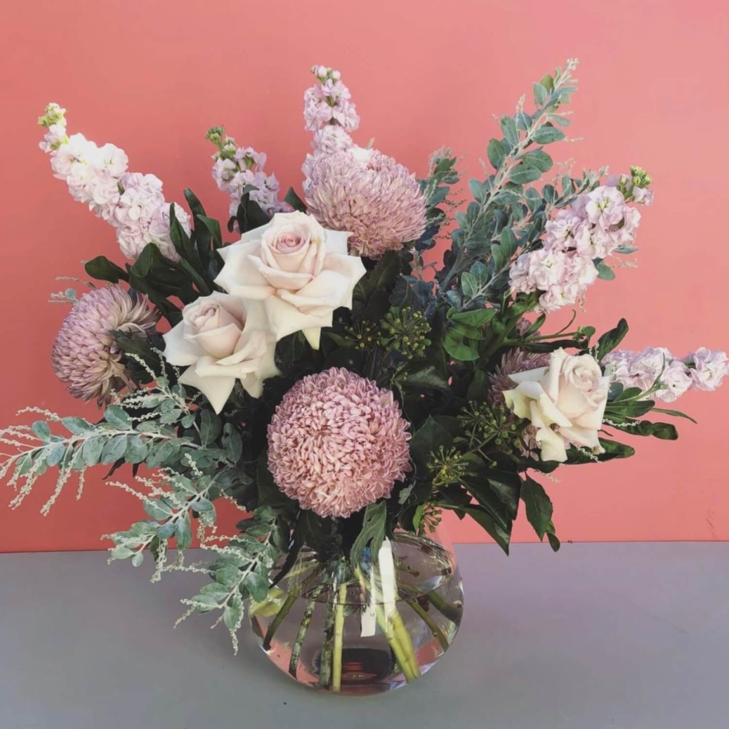 The "Plush Blush" bouquet by FlowersonNortonSt is elegantly displayed in a teardrop vase, featuring pale pink roses, chrysanthemums, and stock flowers. It rests against a pink background with dark leaves and silvery foliage to complete the soft and sophisticated arrangement.