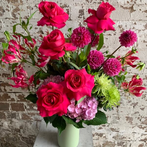 The "Magenta Moments" by FlowersonNortonSt showcases a vibrant bouquet arranged in a ceramic vase, featuring pink roses, dahlias, hydrangeas, and green chrysanthemums. This stunning display of seasonal blooms is beautifully set against a rustic, whitewashed brick wall.