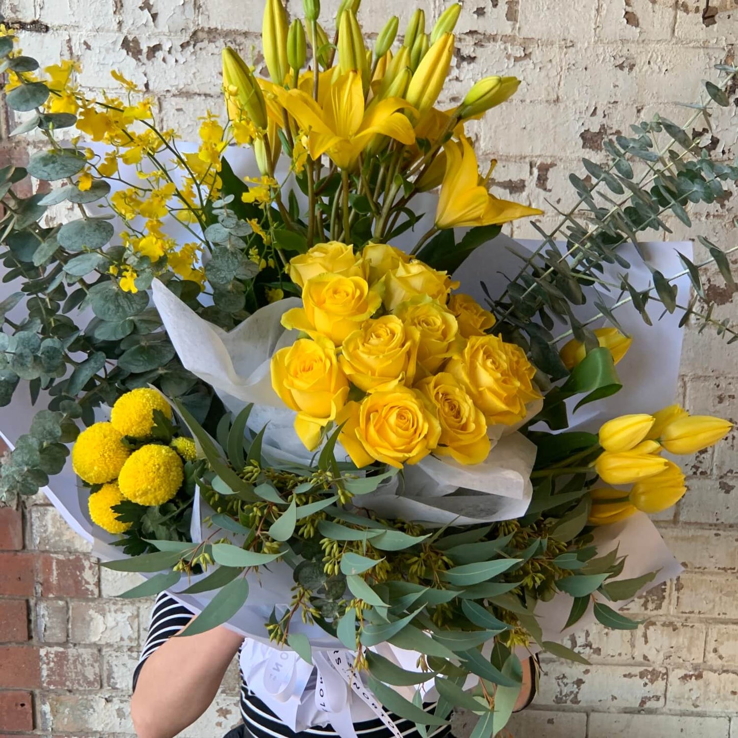 The Colour Me - Yellow Seasonal Bouquet by FlowersonNortonSt features a large mix of yellow roses, lilies, and tulips with eucalyptus leaves, elegantly wrapped in white paper. A person in a striped shirt presents it against a textured brick wall.