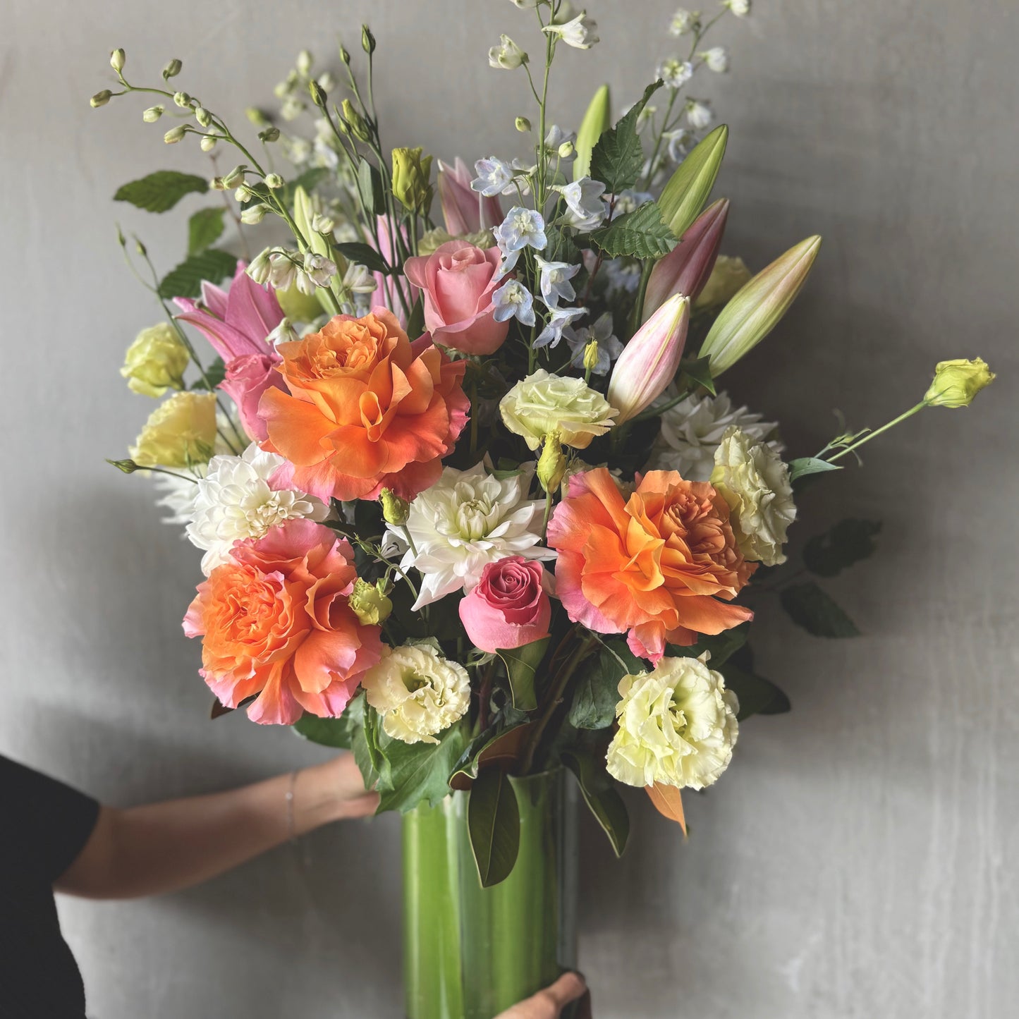 The Pretty Pastels Vase Arrangement by Flowers on Norton St showcases vibrant pink lilies, peach roses, white dahlias, and green leaves. It brightens any day with its charming floral display against a neutral gray backdrop, held by a person in a black shirt.