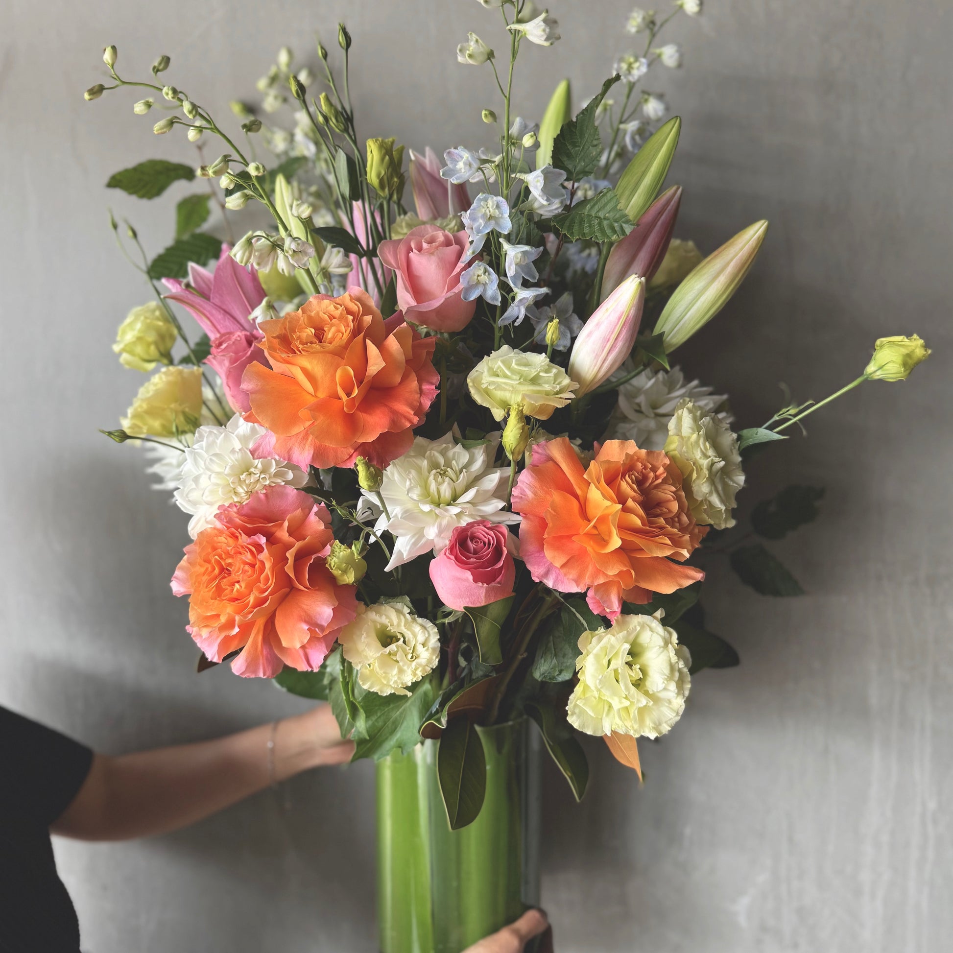 The Pretty Pastels Vase Arrangement by Flowers on Norton St showcases vibrant pink lilies, peach roses, white dahlias, and green leaves. It brightens any day with its charming floral display against a neutral gray backdrop, held by a person in a black shirt.