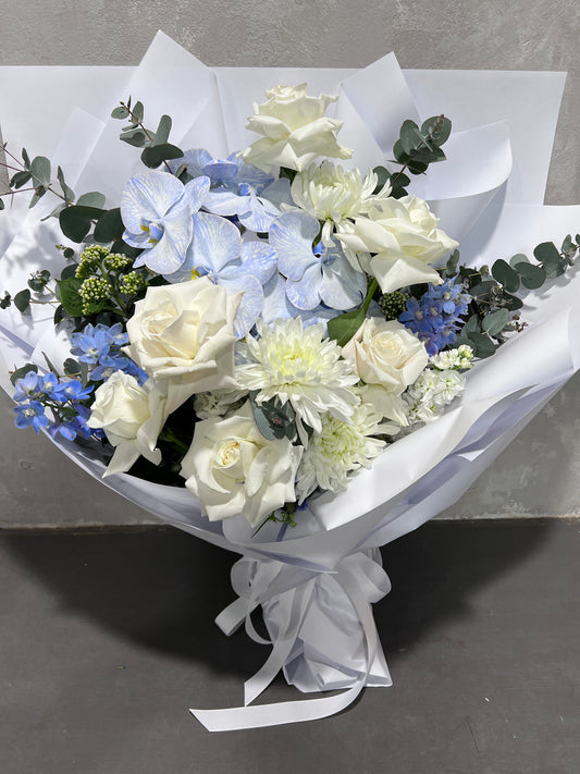 The Blue Daze Seasonal Bouquet by FlowersonNortonSt, showcasing a striking combination of white roses, chrysanthemums, and phalaenopsis orchids elegantly wrapped in white paper and tied with a delicate ribbon, rests gracefully on a gray surface.