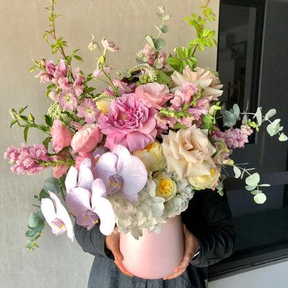 A person holds a vibrant bouquet in the Fairy Garden | March Pink Vase by Flowers on Norton St. The arrangement features pink roses, white orchids, yellow roses, and greenery against a neutral background, showcasing Emma's signature style with this colorful display.