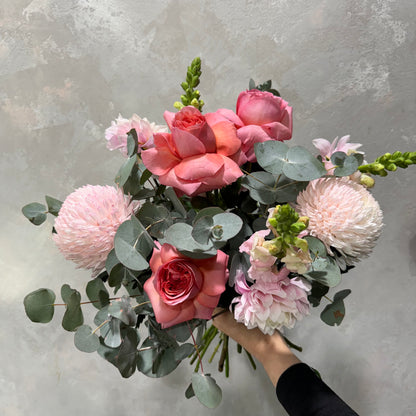 A person holding the "Pretty in Pink Mixed Flowers" bouquet from Flowers on Norton St, featuring pink roses, pale pink chrysanthemums, and eucalyptus leaves against a textured gray background.