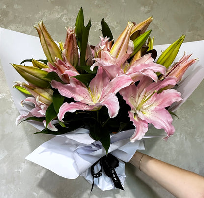 A bouquet of Casablanca Lily flowers by FlowersonNortonSt, featuring pink Oriental lilies and green leaves, is elegantly wrapped in white paper and tied with a black ribbon. A person's hand presents these long-lasting flowers against a simple light-colored background.