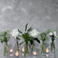A collection of vases, products from the Style the Table Kit - Mother's Day Edition by Flowers on Norton St, elegantly displays fresh flowers such as white roses and green foliage against a textured gray wall. Nearby, small glass candle holders with tea lights enhance the ambience of the table setting, creating a peaceful and refined scene ideal for celebrating Mother's Day.