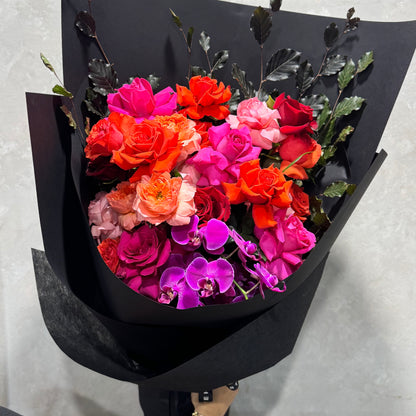 The Colombian Rose Extravaganza Arrangement by Flowers on Norton St is a vibrant mix of red, orange, pink, and fuchsia Colombian roses with bright purple Phalaenopsis orchids and dark foliage, elegantly wrapped in black paper.